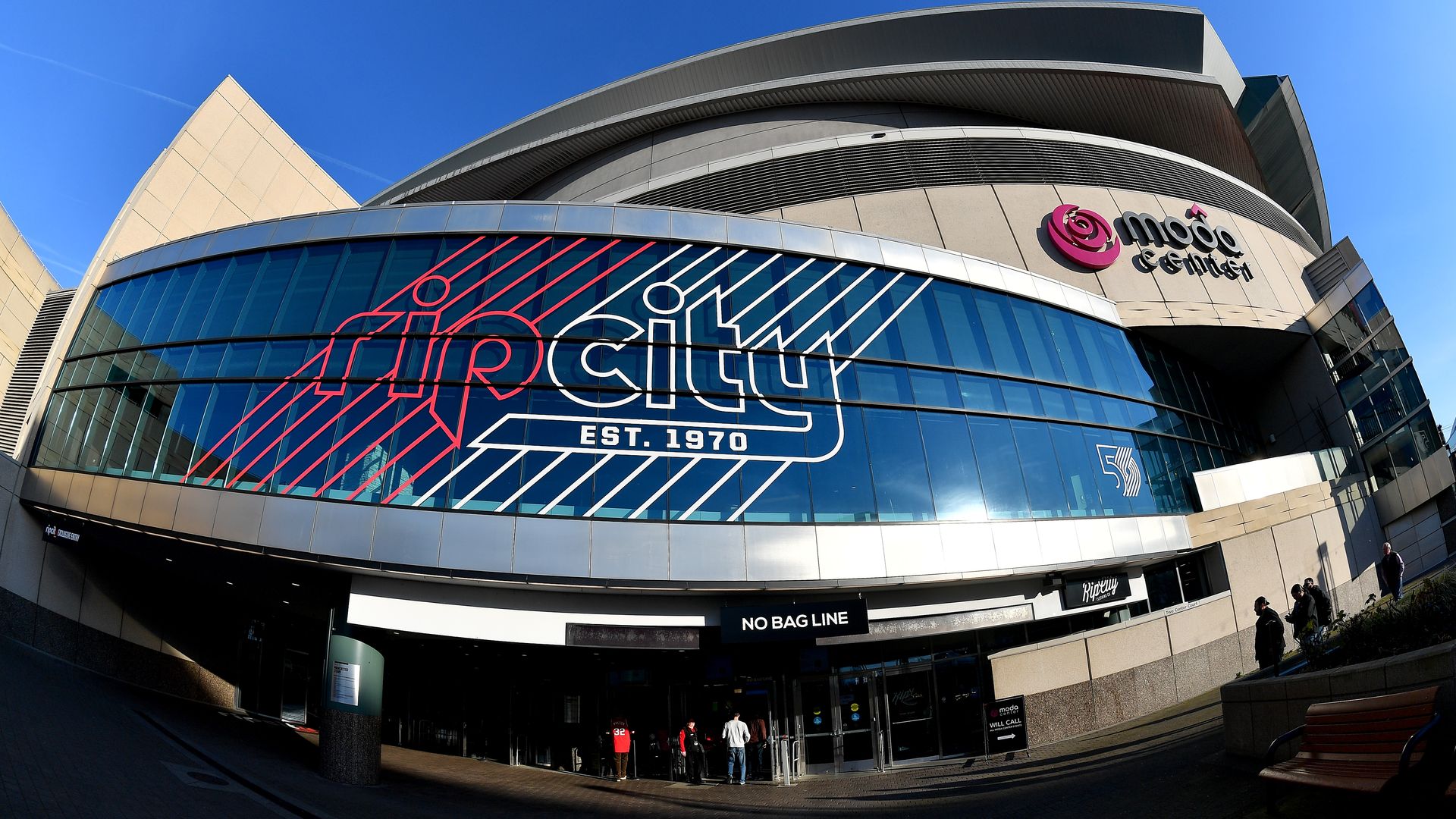 Exterior of the Moda Center arena with large Rip City EST. 1970 graphics on blue tinted glass windows, a few people near the entrance, and clear blue sky above.