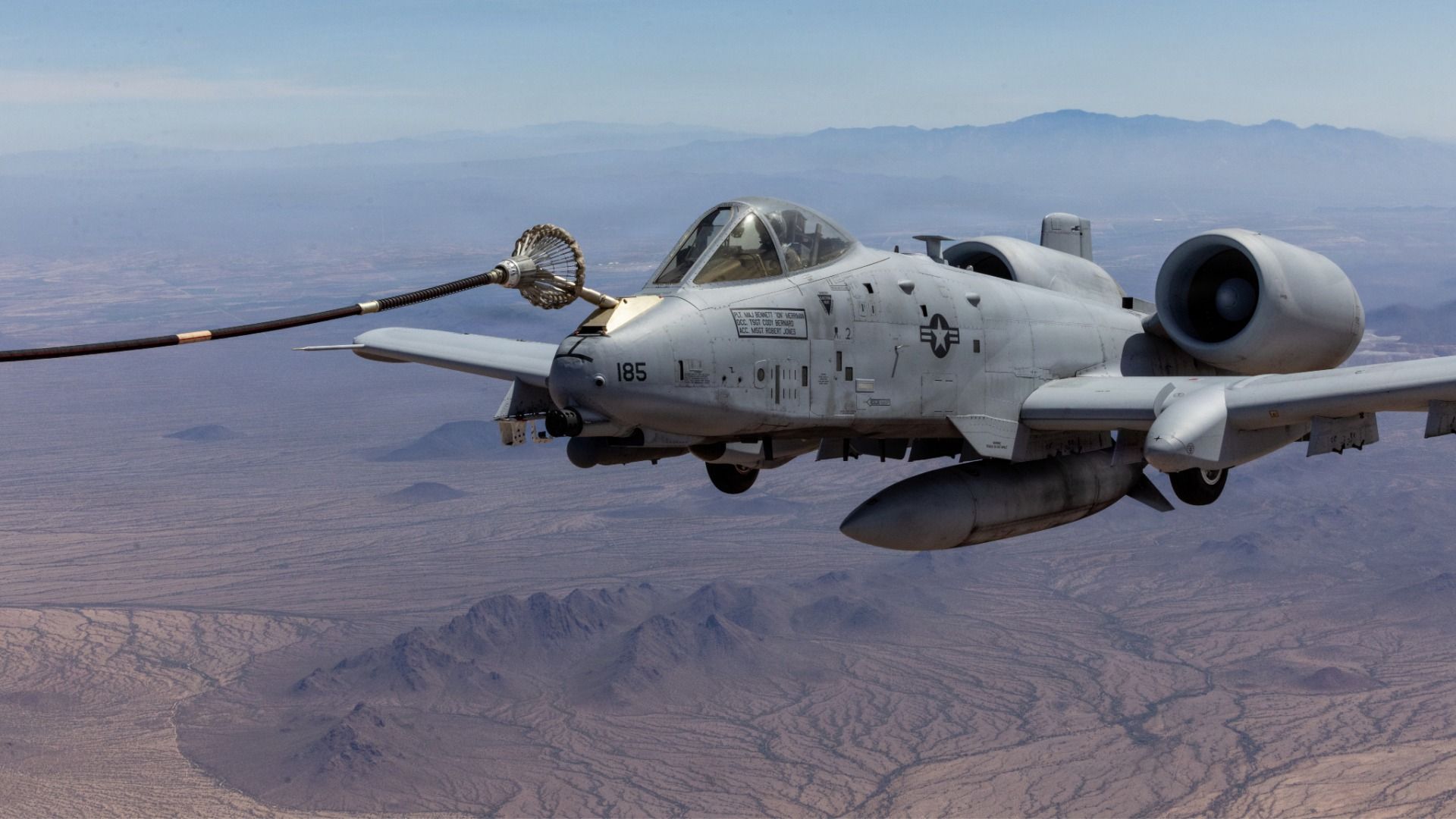 Gray A-10 aircraft in flight over a desert with distant mountains. A long nose refueling probe reaches toward a drogue.