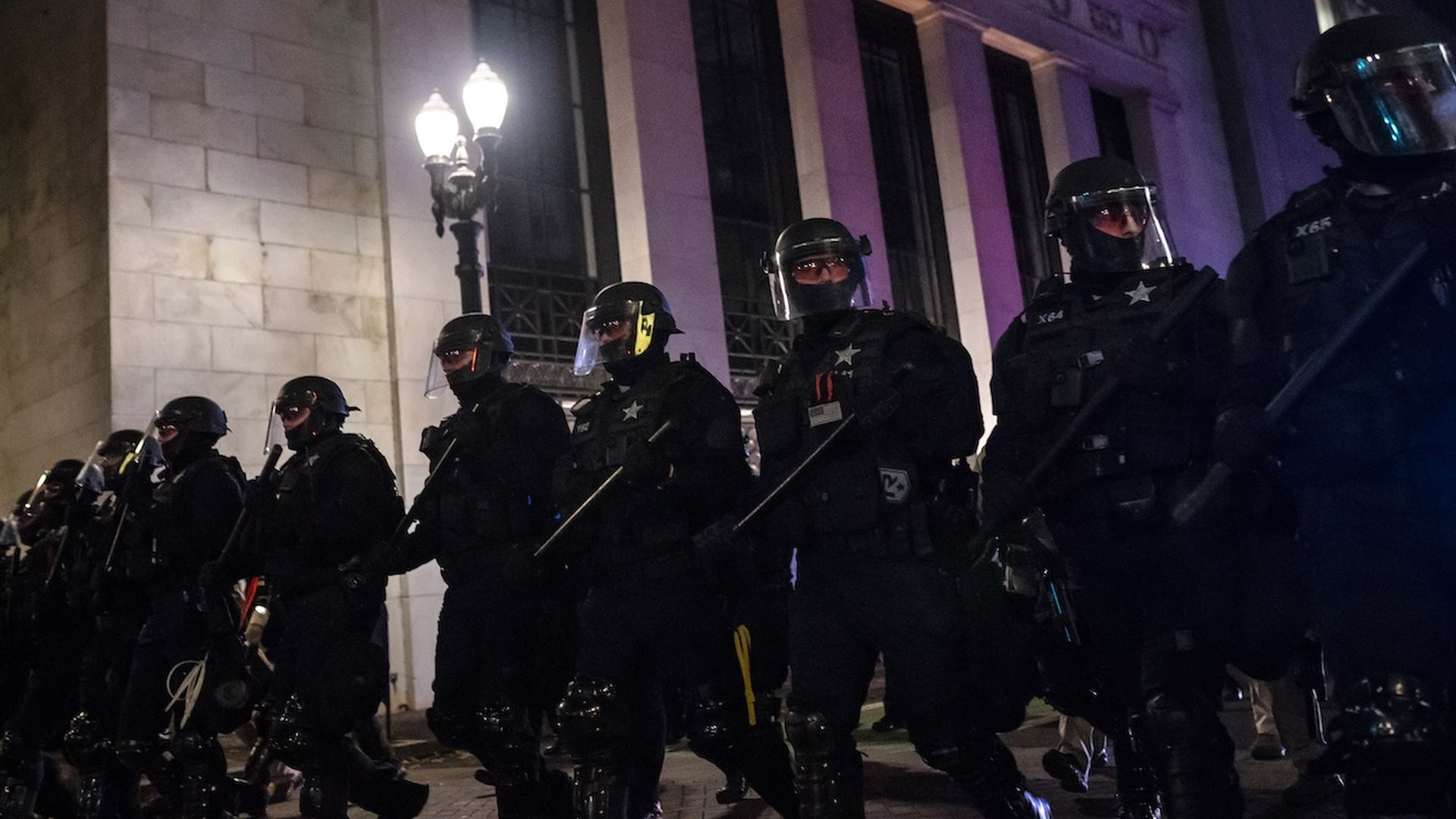 A photo of a group of police officers dressed in protective riot gear in a single line in front of a white marble column building.