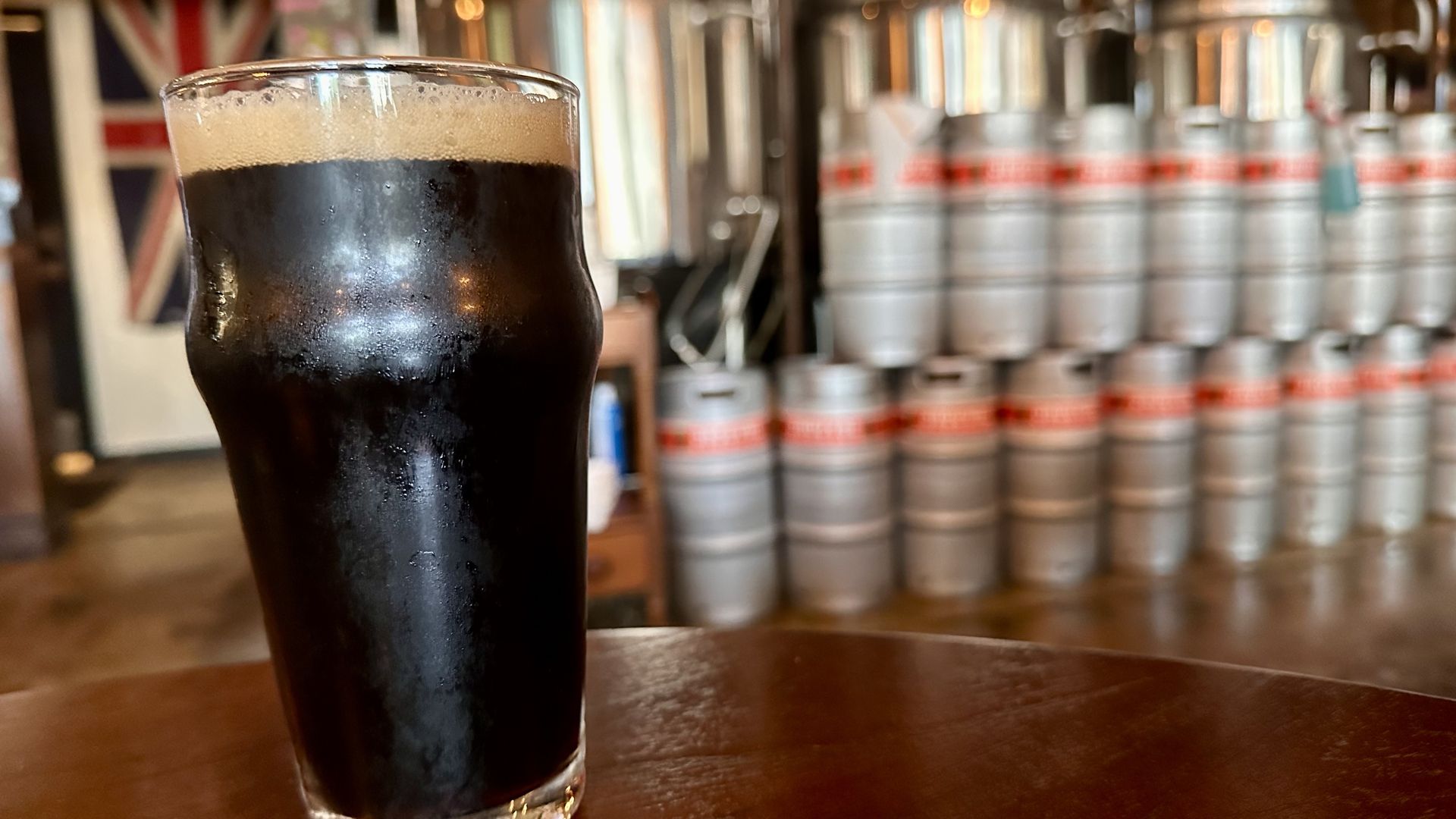 A pint glass filled with dark beer with frothy head sits on a wooden table inside a brewery; metal kegs stacked in rows can be seen blurred in the background.