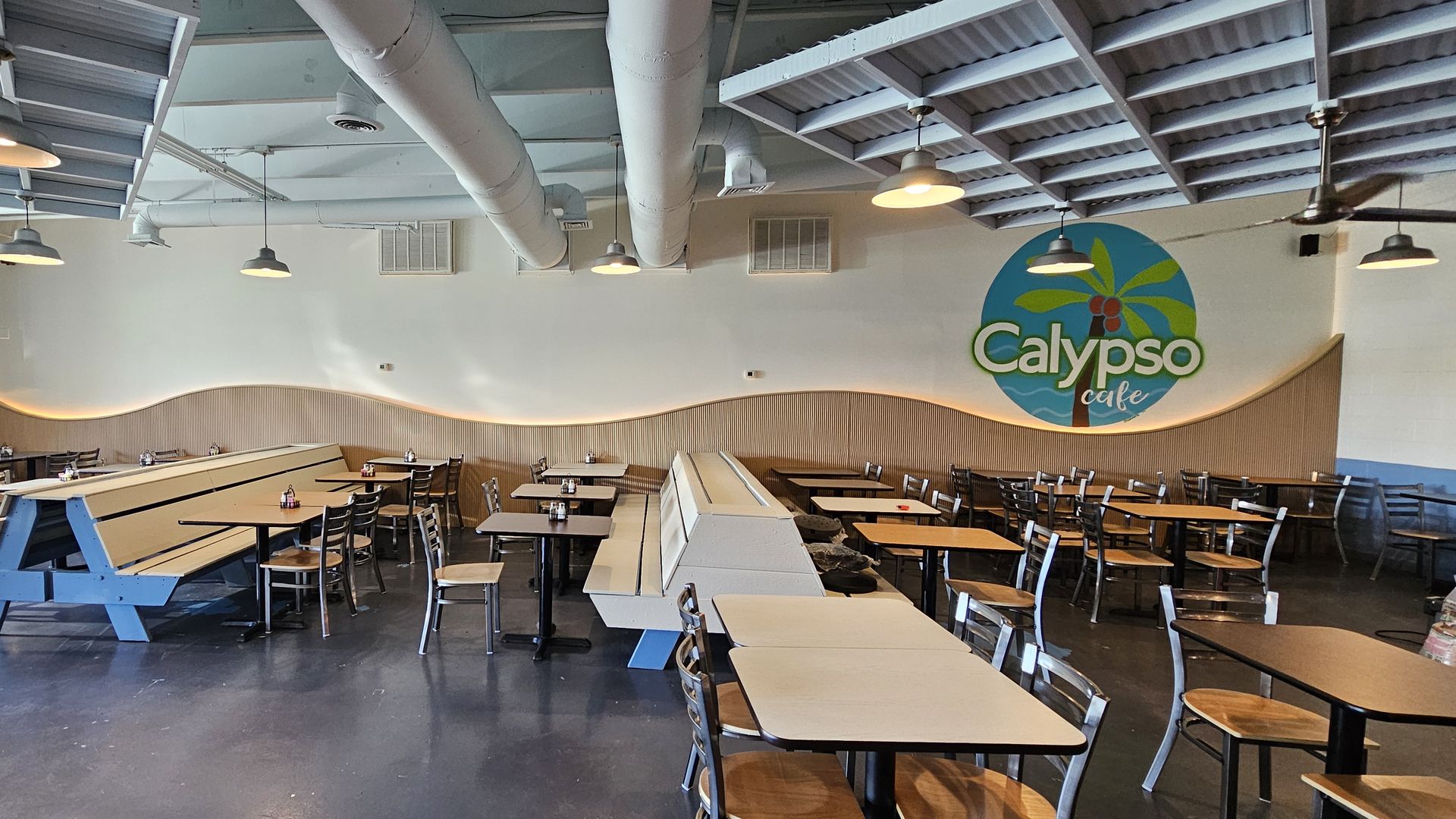 Bright cafe interior with exposed white ducts and a gray ceiling. Pendant lights illuminate wood tables and metal chairs; a turquoise circular Calypso cafe logo sits on the right wall above paneling.