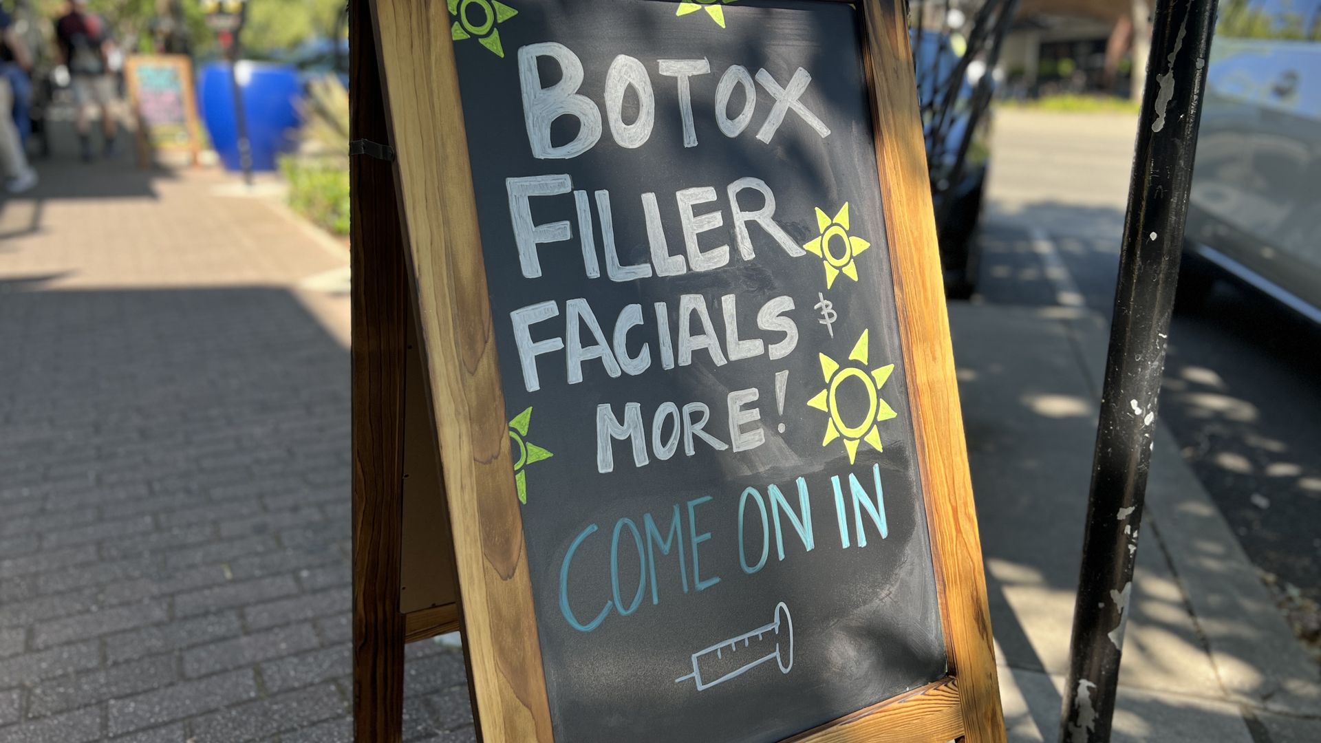 Outdoor chalkboard sign in a wooden frame on a sunny sidewalk, white bold letters with yellow sun doodles advertising fillers and facials