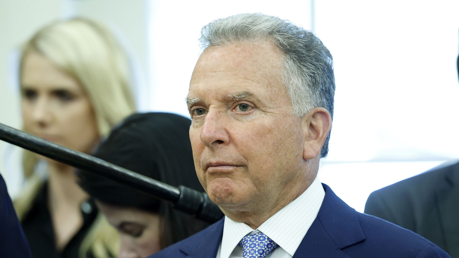 Older man with gray hair in a blue suit and patterned blue tie at a formal event, with blurred people and a microphone in the background.