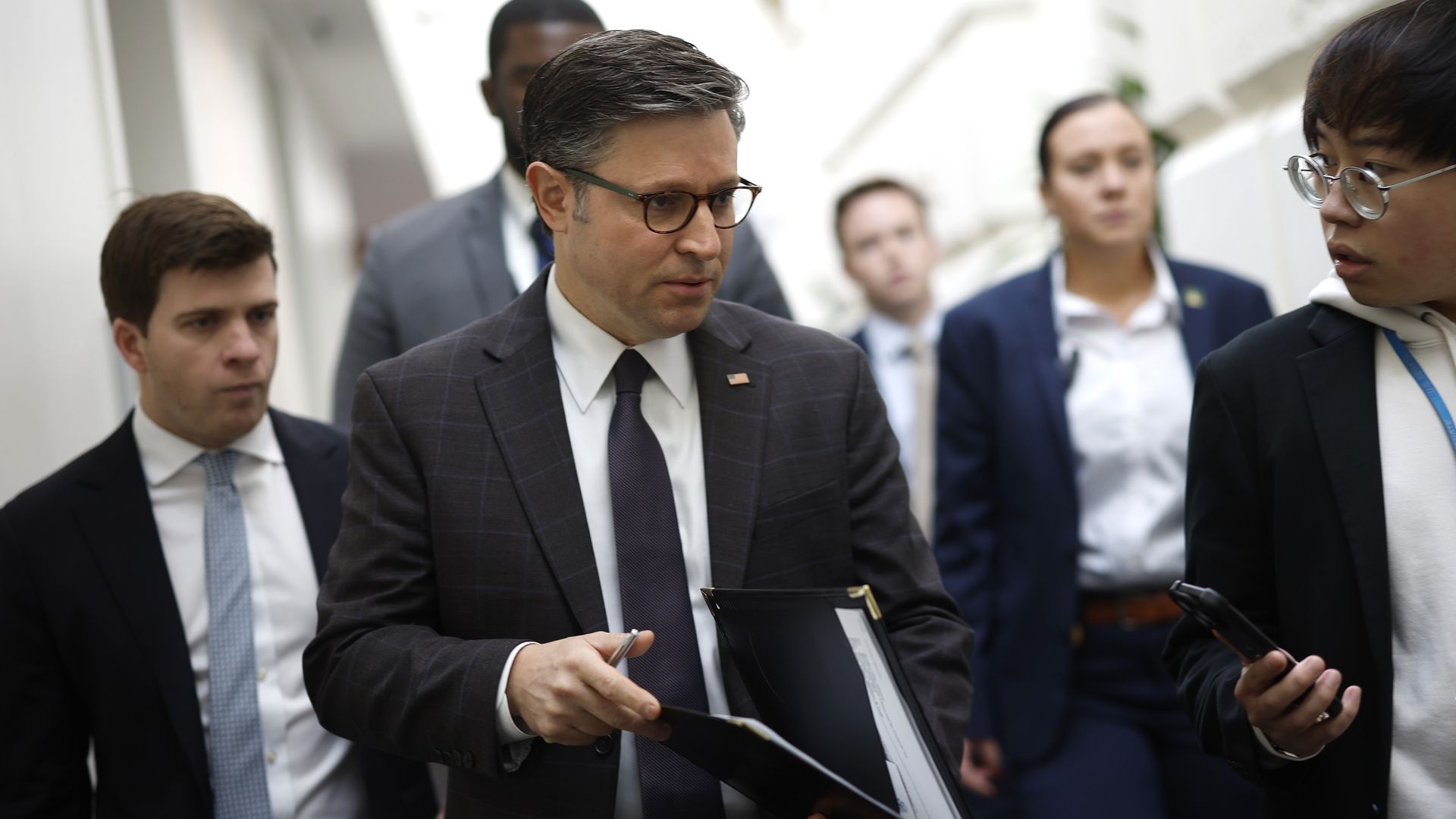 House Speaker Mike Johnson, wearing a gray suit and walking through a white hallway while surrounded by aides, security and press.