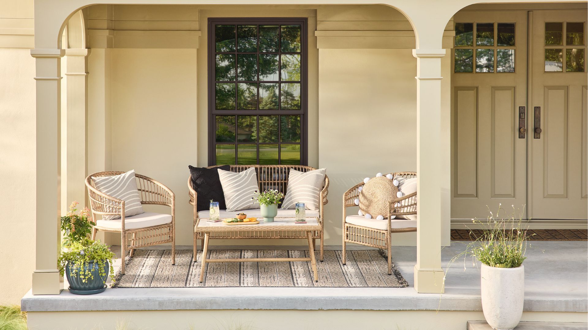 A front porch setup featuring a wicker furniture set with pillows, a rug and refreshments on a table.