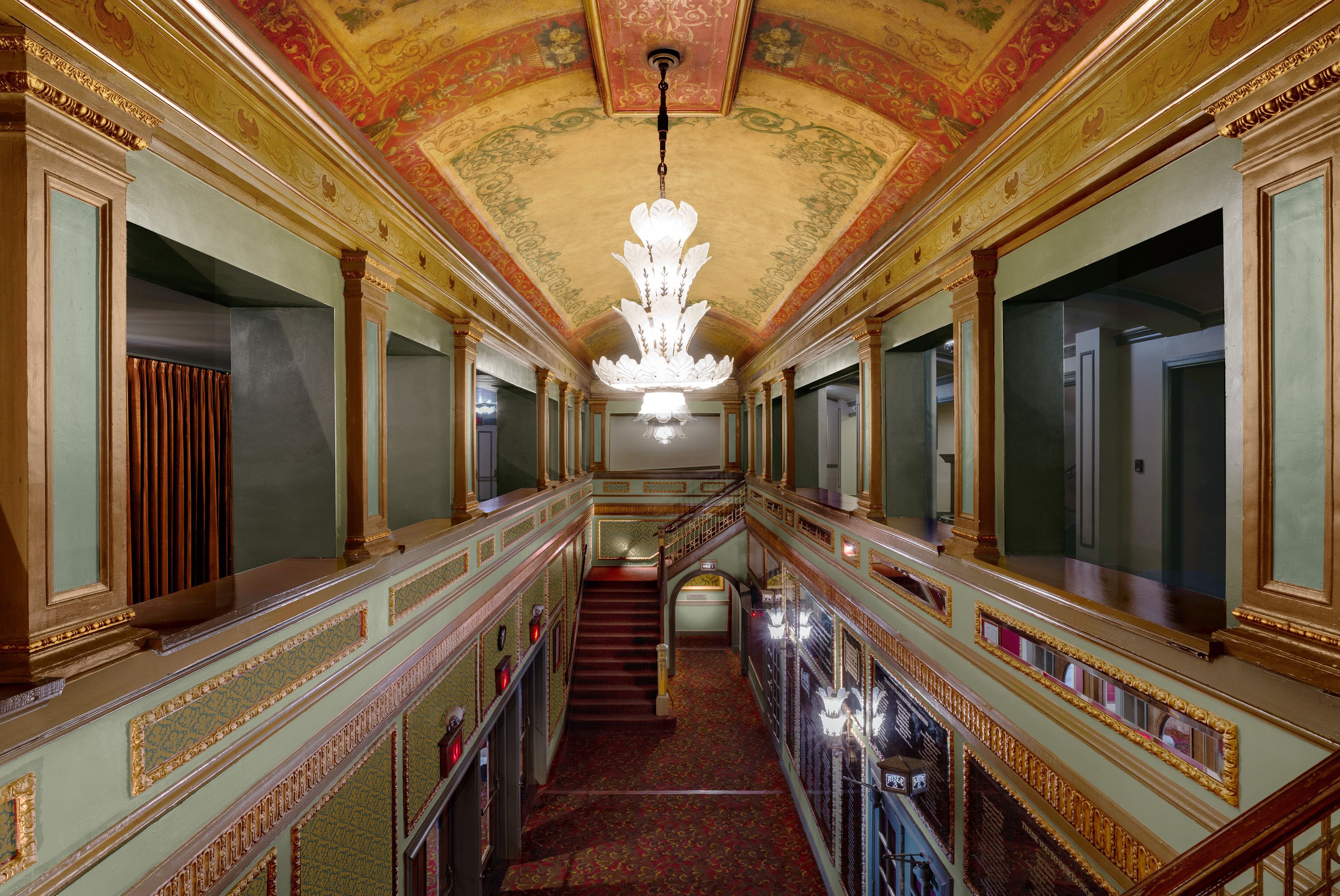 Elegant hotel corridor with green walls and gold trim, ornate gold-accented arches on both sides, a tall white leaf-shaped chandelier hanging from a painted ceiling, and a red patterned carpet.