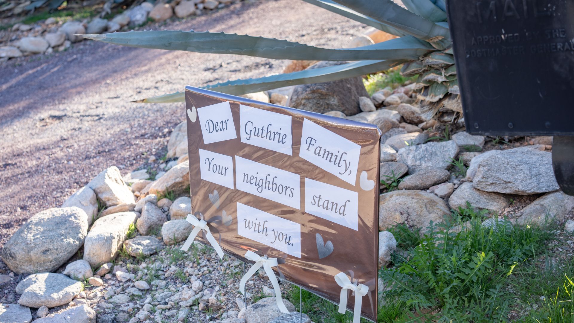 Brown sign with white and silver hearts and text reading "Dear Guthrie Family, Your neighbors stand with you," displayed outdoors among rocks and plants.