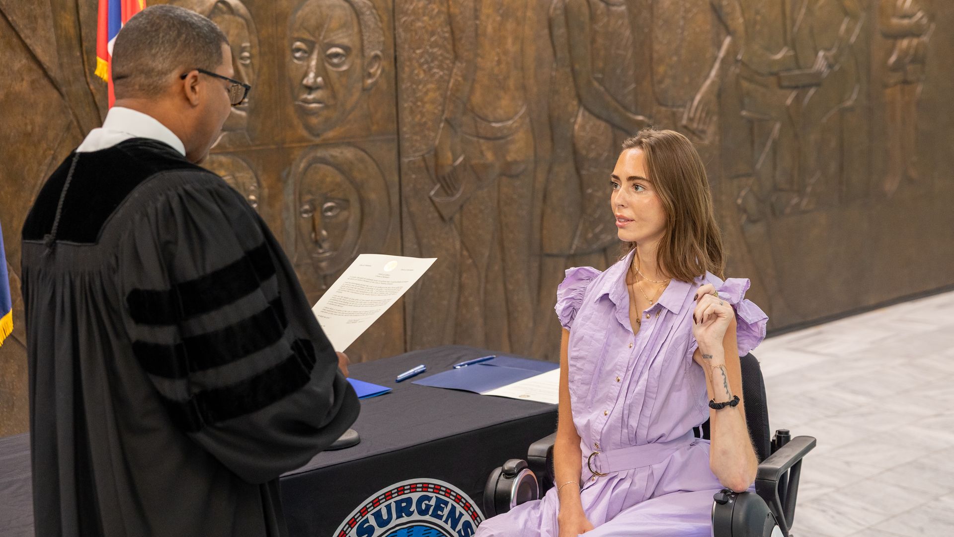 A man in a black robe reads a document to a woman in a lavender dress sitting in a wheelchair, against a bronze wall with faces and figures carved in it.