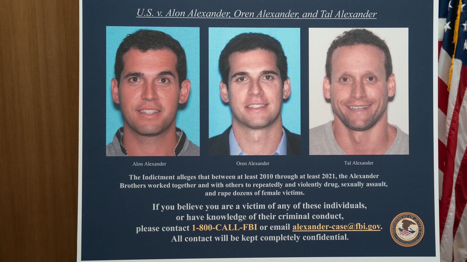 Poster board with three mugshots on blue panels, a large indictment text block, and an American flag visible to the right.