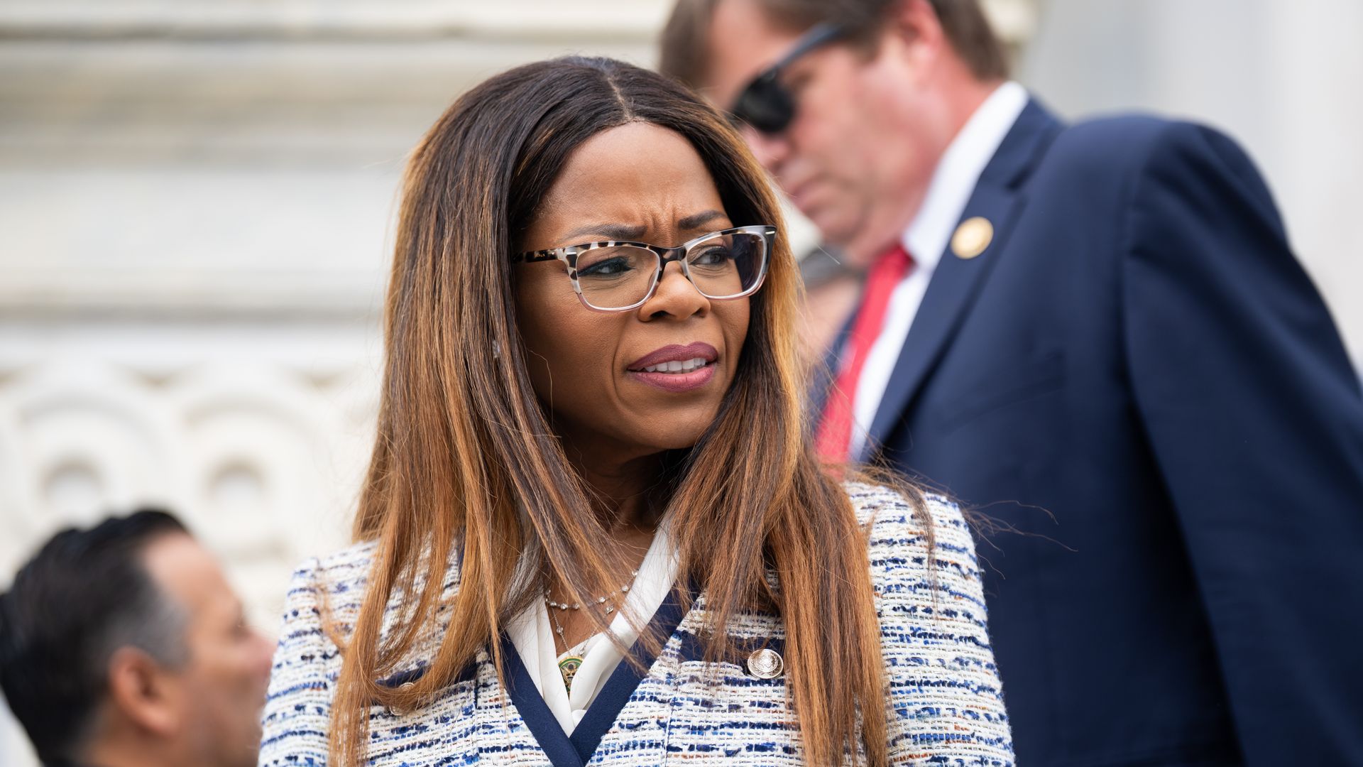 Woman with long brown hair and glasses wearing a white and blue checked jacket stands outdoors with two blurred people in the background, one in a dark suit and red tie.