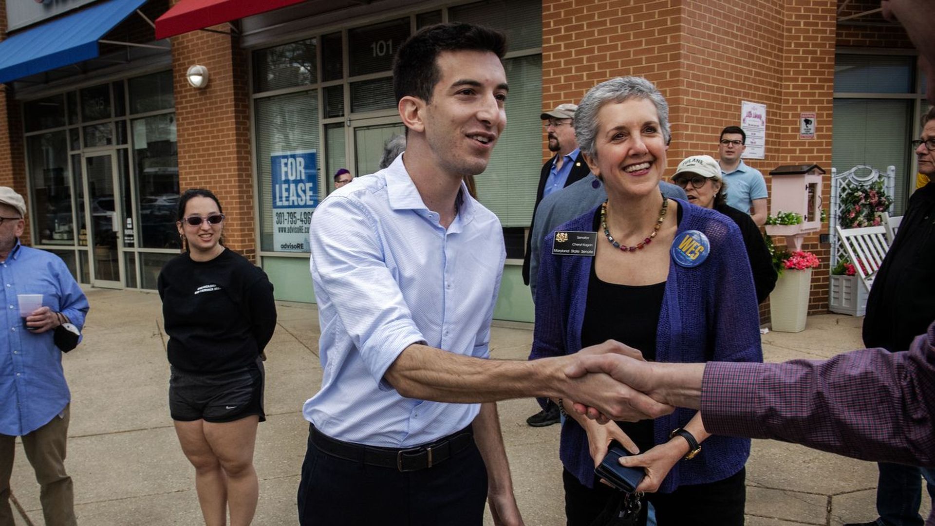 Joe Vogel shakes the hand of someone who is not shown in the picture while campaigning for office. A woman smiles next to him.