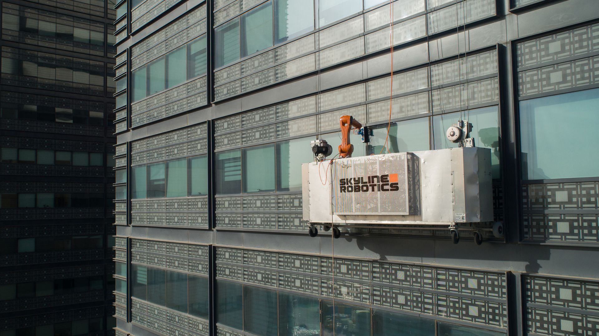 A window-washing basket on the side of a skyscraper with robots instead of humans in it.