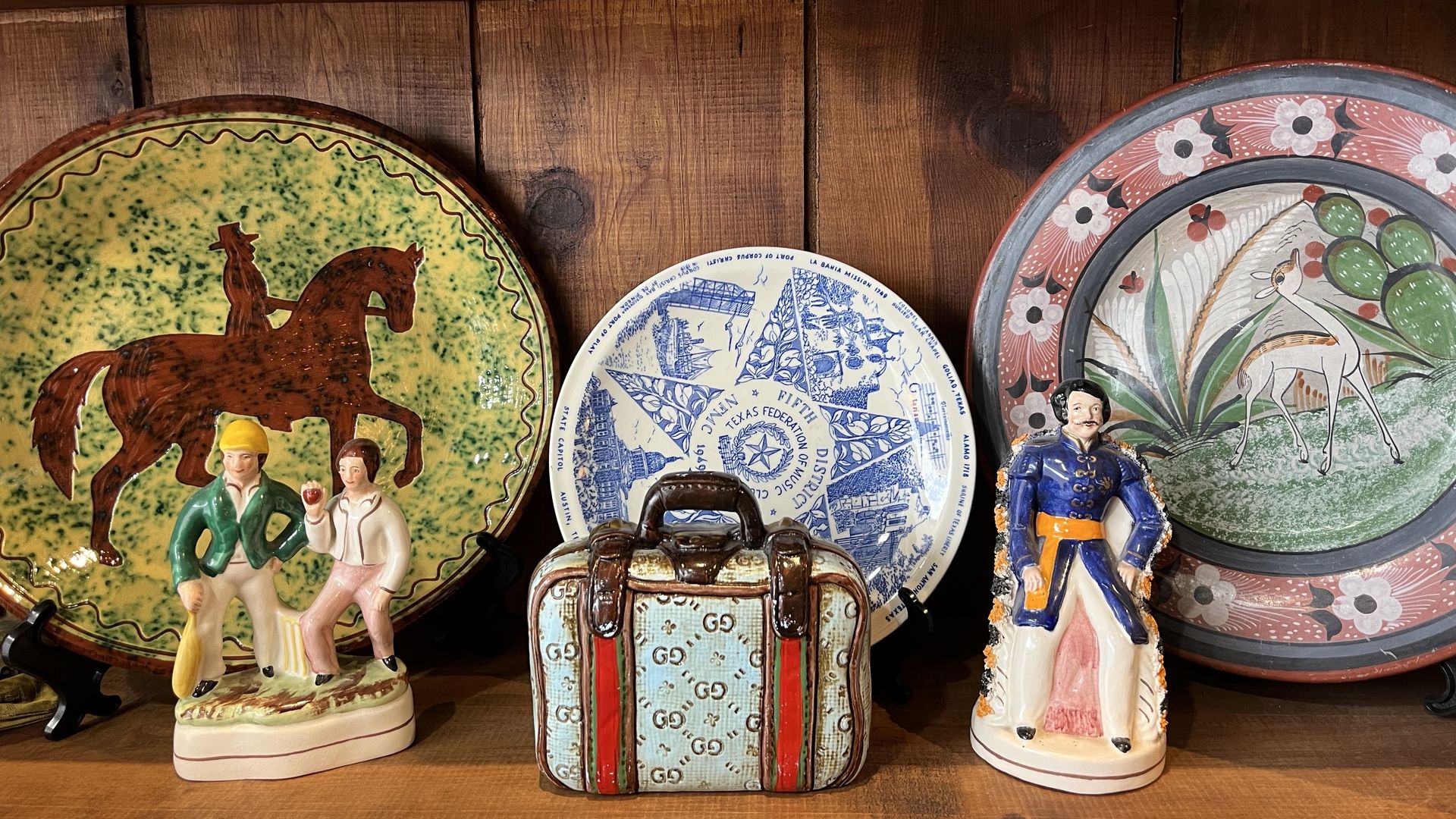 Wooden shelf with assorted ceramics: a large green plate with a brown horse and two figures, a blue-and-white plate with travel motifs, a pink floral plate, and a decorative mini trunk.