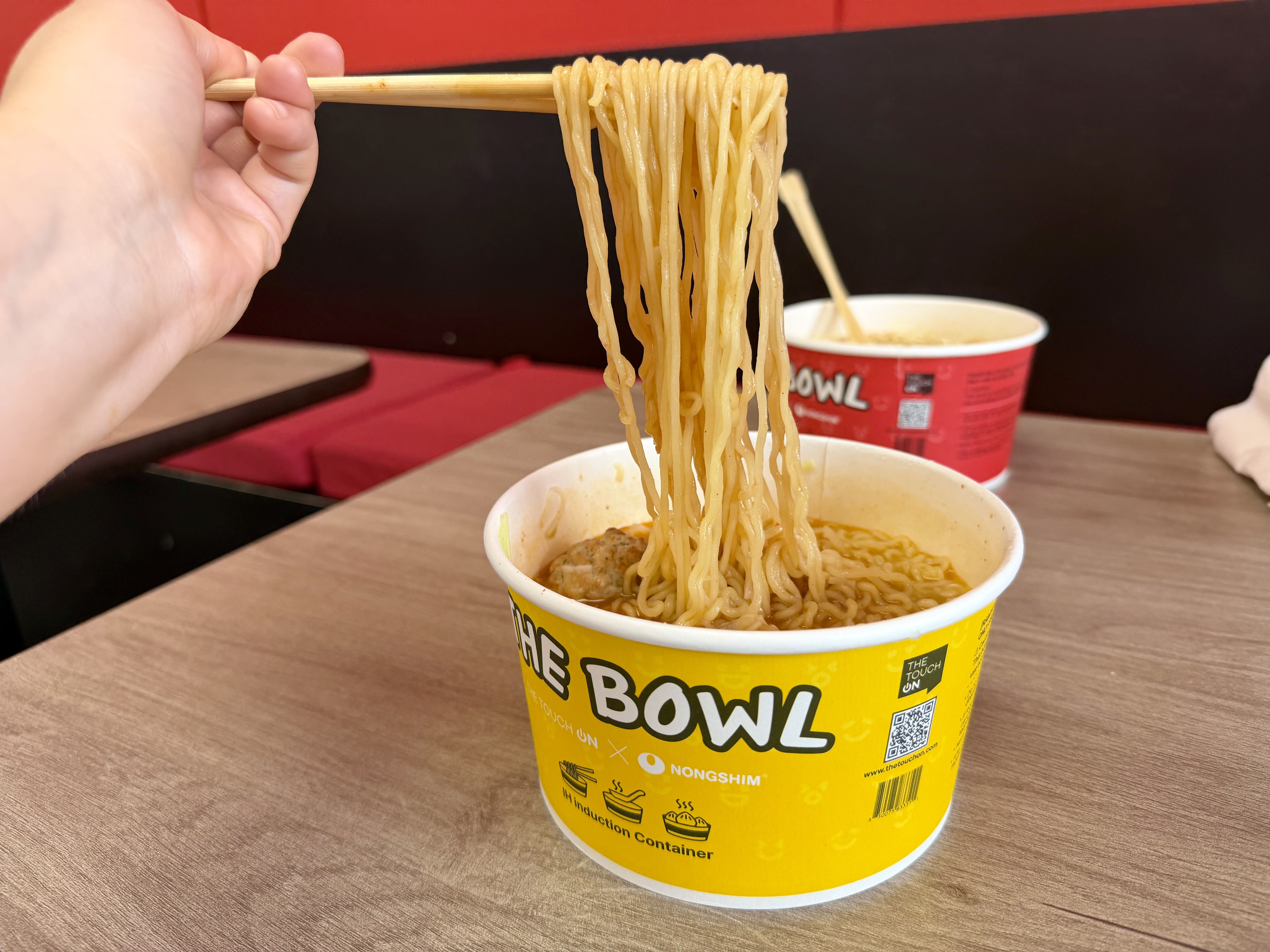 Close-up of a hand using wooden chopsticks to lift yellow ramen noodles from a bright yellow cup labeled BOWL; broth visible and a red container is in the background.