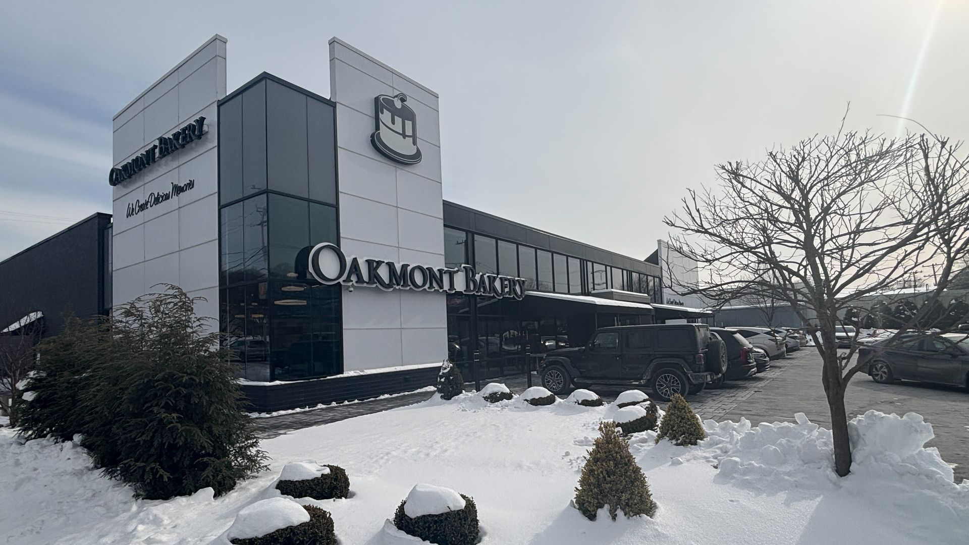 Oakmont Bakery building in winter with snow-covered bushes, bare tree, and parked cars under gray sky and soft sunlight.