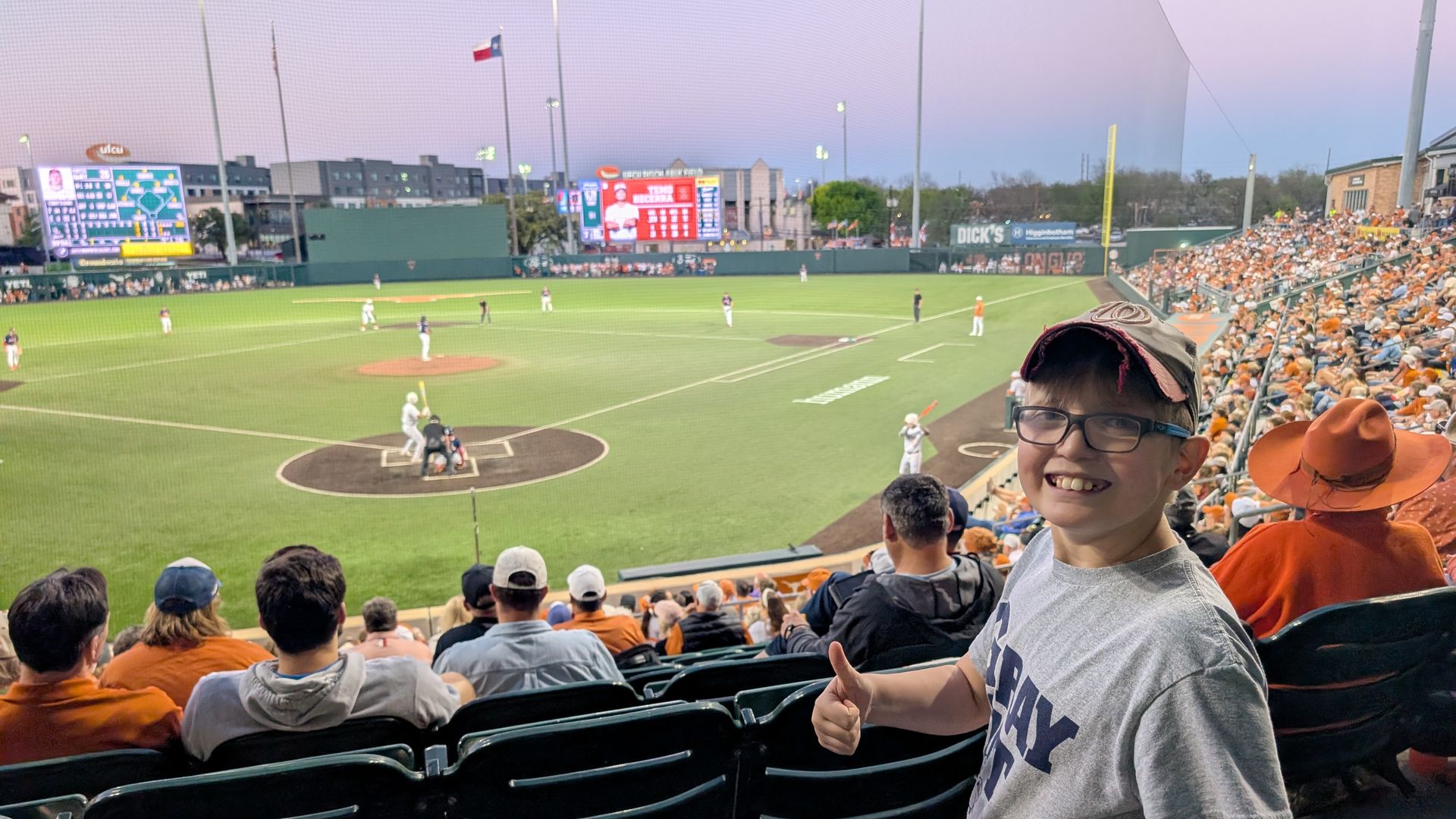 Smiling boy with glasses in a gray shirt and cap gives a thumbs-up from orange-clad stands as a baseball game unfolds on a green field with players and a scoreboard in the background.