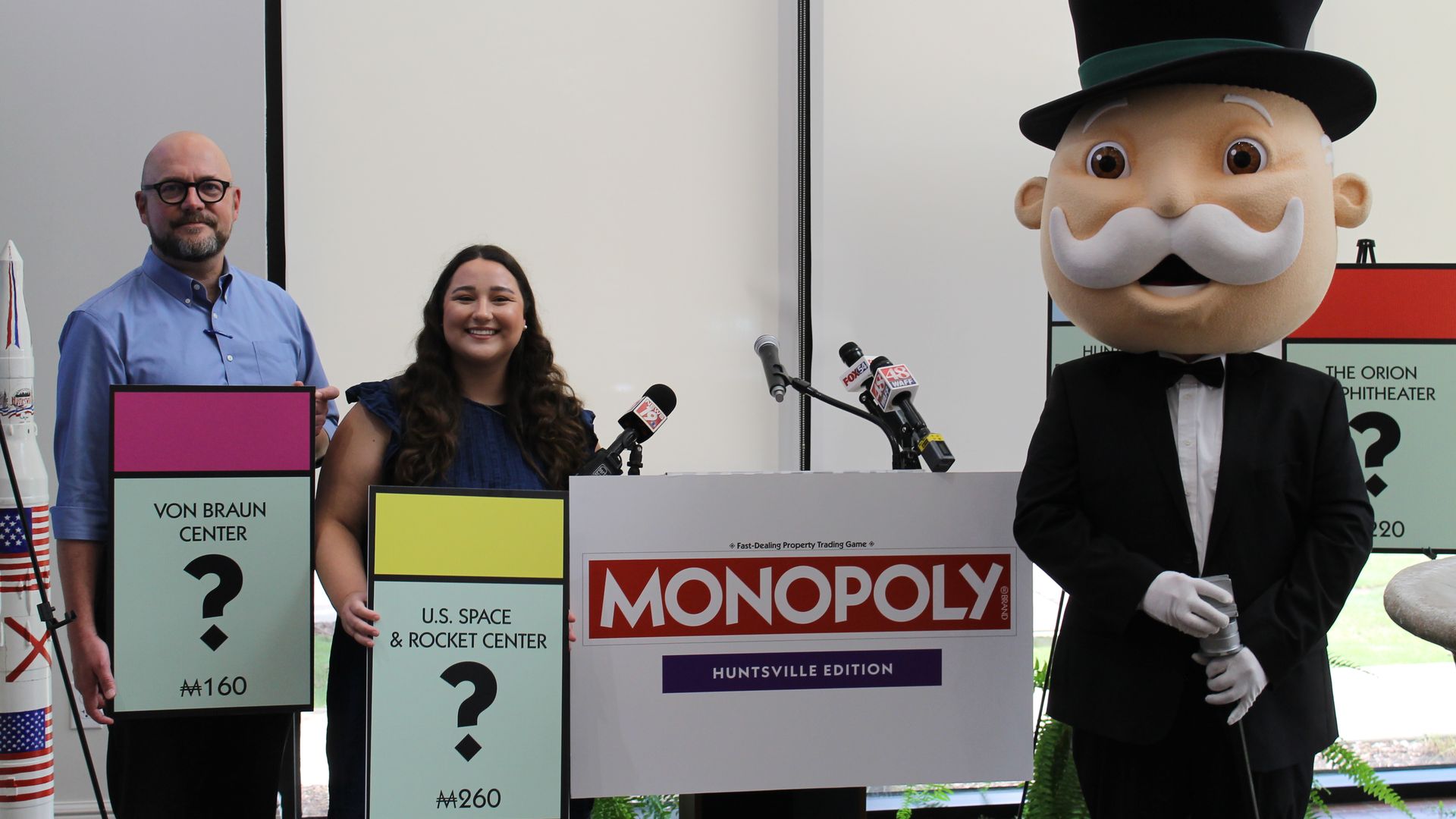 Press event with three people and a large Mr. Monopoly mascot; left man in blue shirt and glasses, center smiling woman, right mascot in a tux; oversized Monopoly boards and the logo visible.