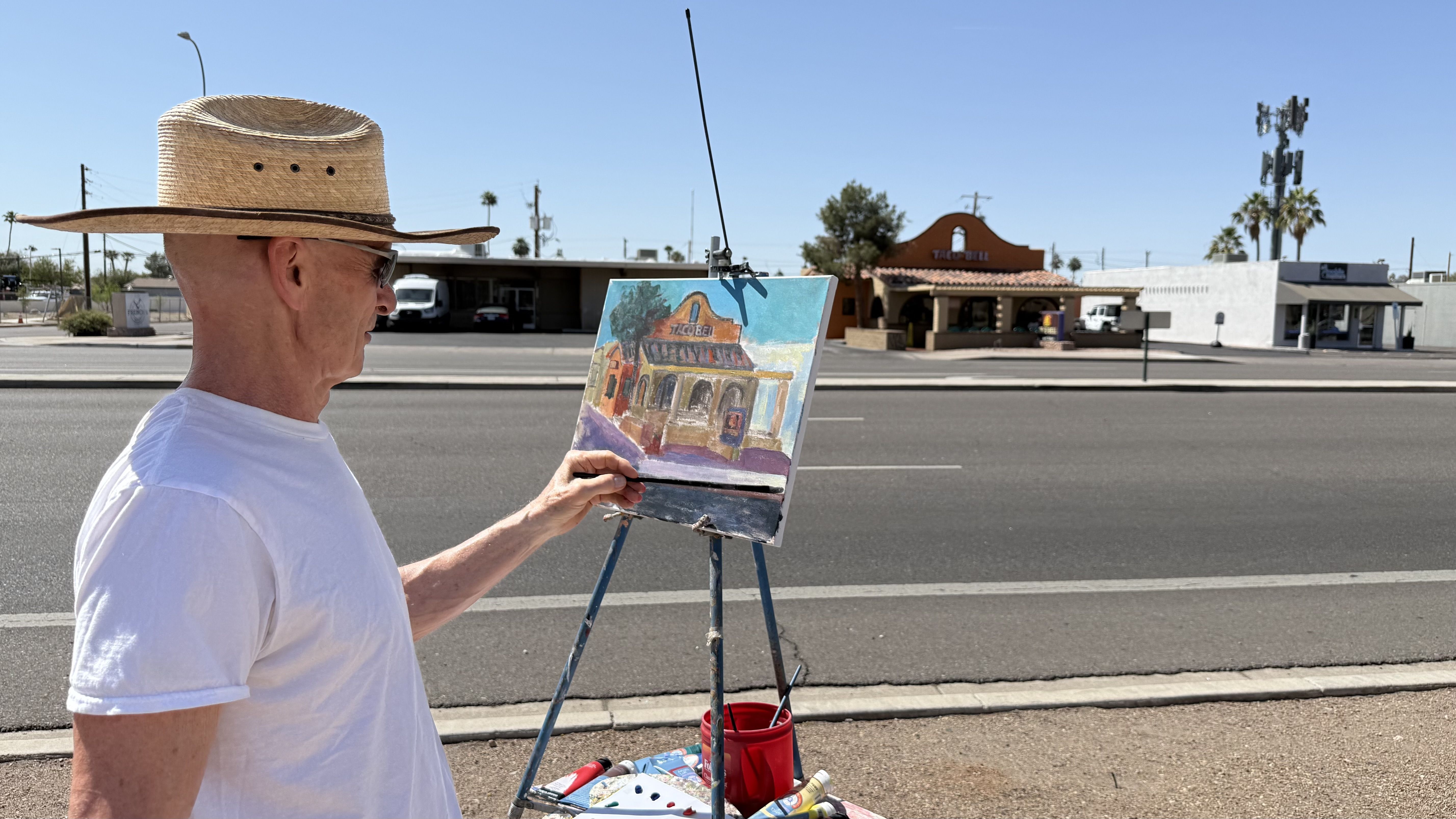 A man in a white t-shirt and a broad-brimmed hat paints a picture of a mission-style Taco Bell from across the street.