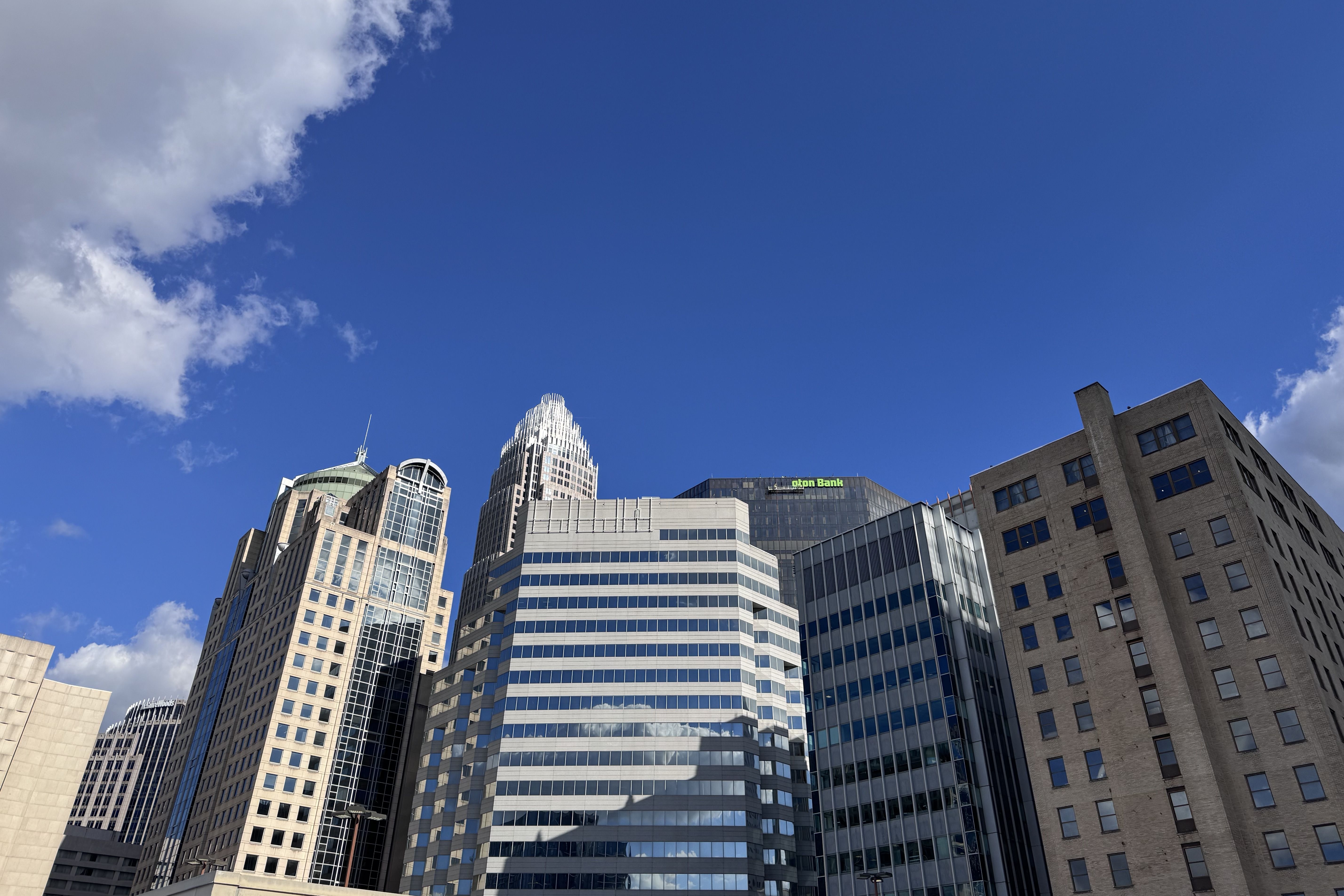 Blue sky over a cluster of modern skyscrapers with glass and brick facades; a tall tower with a pointed crown is center, and a green bank sign sits on a nearby building.