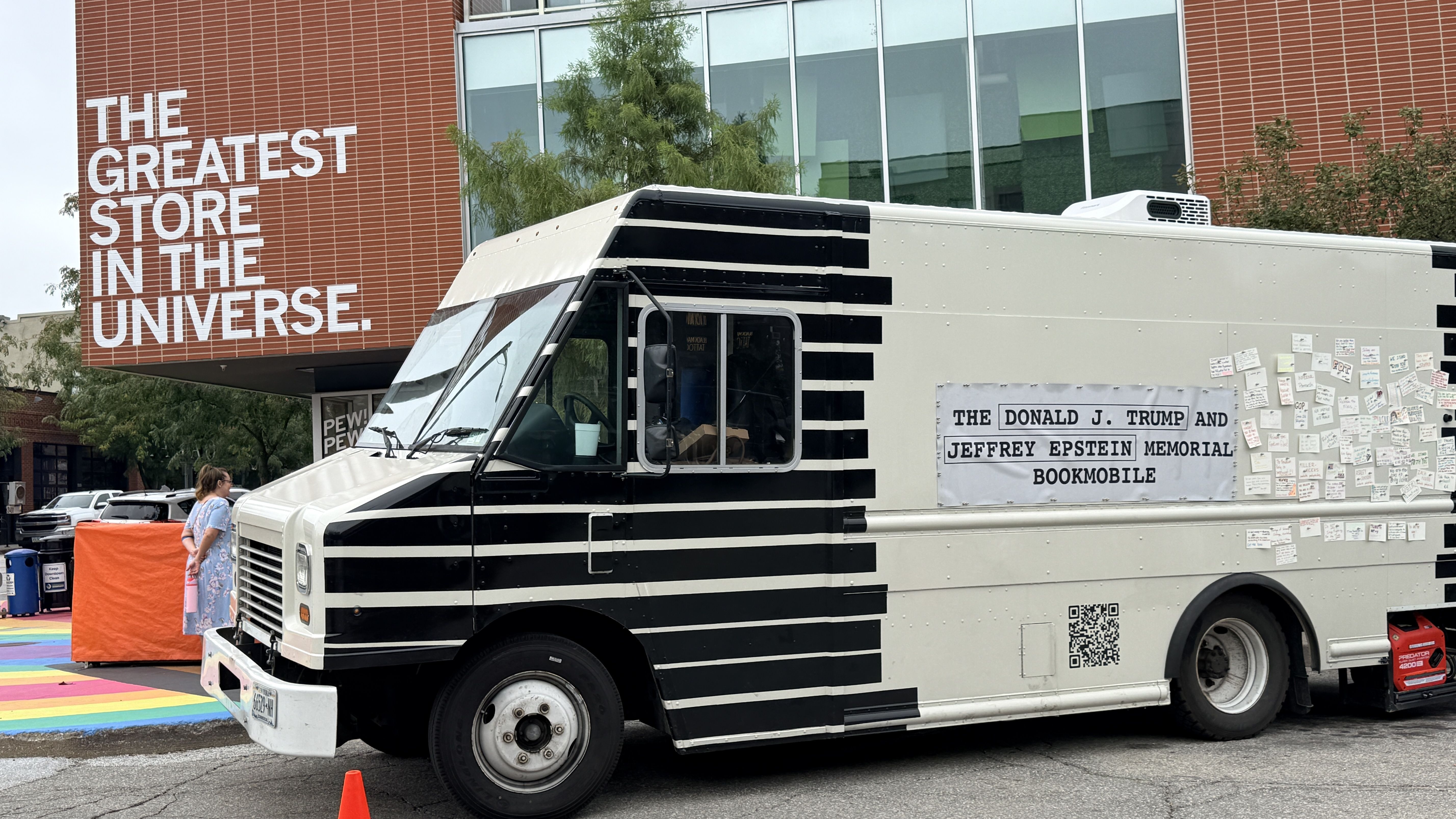 White and black striped bookmobile beside a brick building with a large sign reading "THE GREATEST STORE IN THE UNIVERSE"; numerous notes are posted along the vehicle's side.