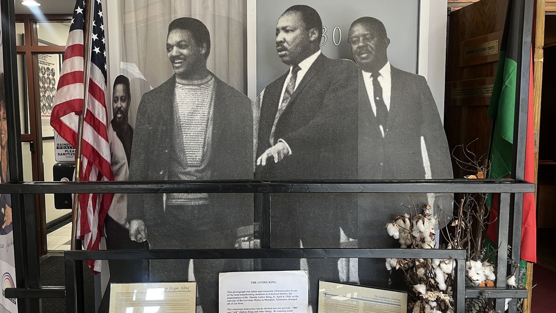 Black-and-white photo collage of four men, including Martin Luther King Jr. in a suit, with an American flag and informational plaques below them in a display.