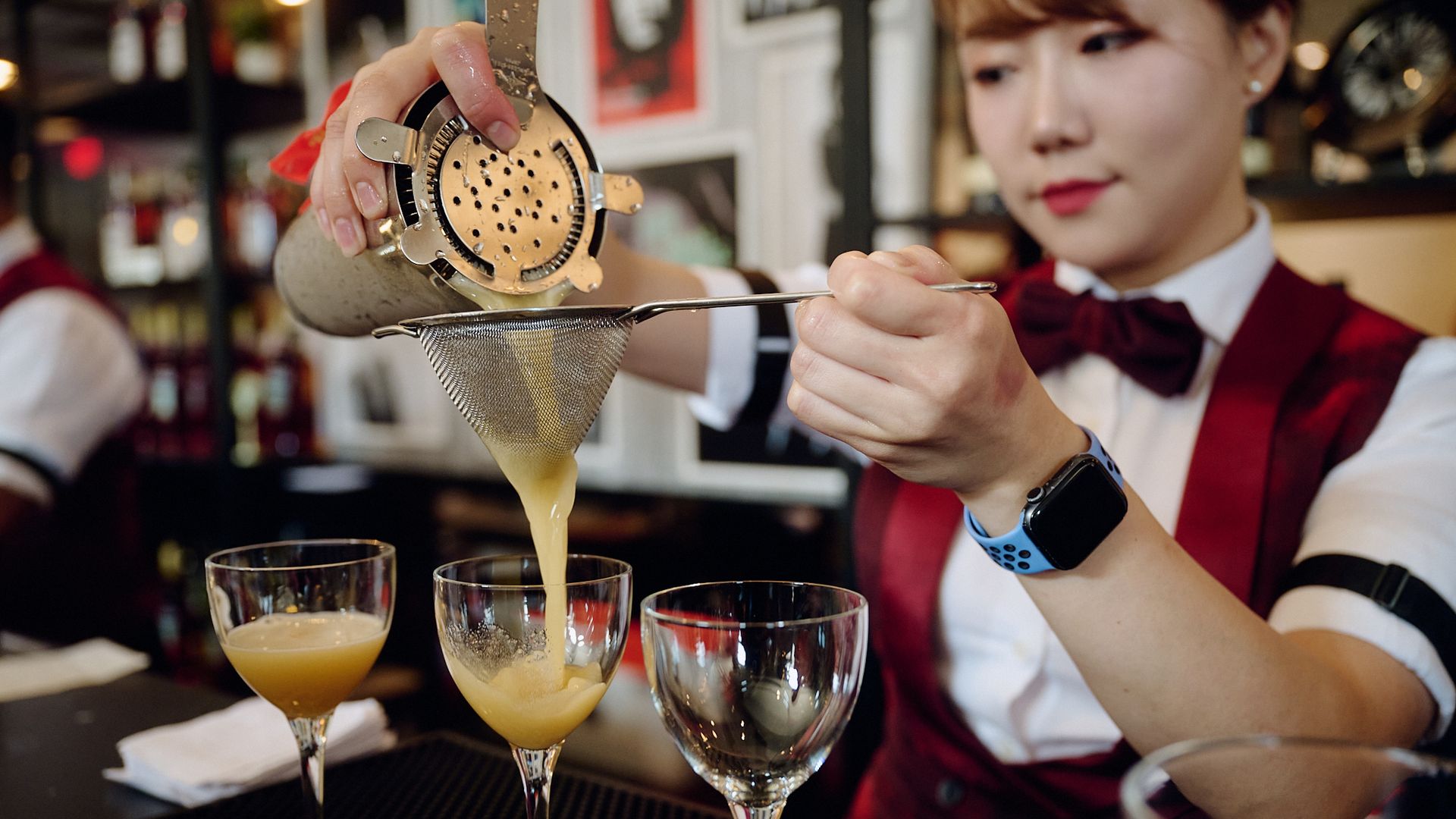 A bartender pours a drink from a shaker through a strainer and into one of three glasses lined up on a bar.