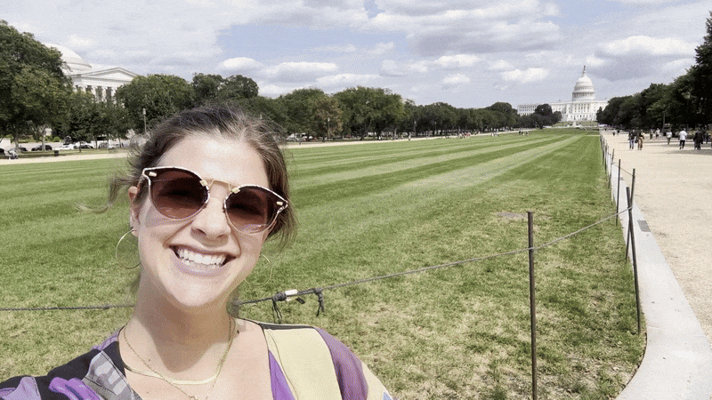 A woman wearing sunglasses and a patchwork jacket takes a selfie video pointing at the U.S. Capitol.