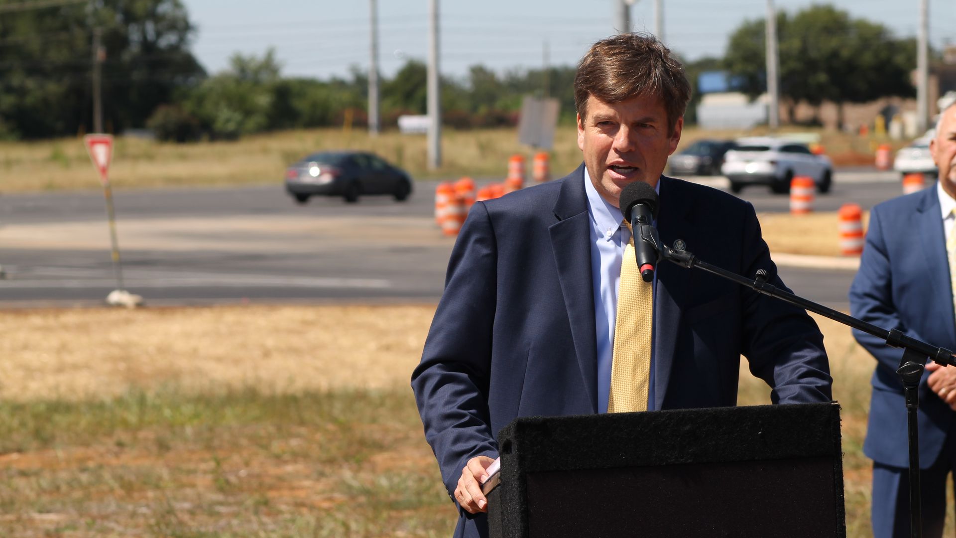 Man in navy suit and yellow tie speaking at outdoor podium with microphone, traffic lights and cones in background, other man stands behind him in blue suit.