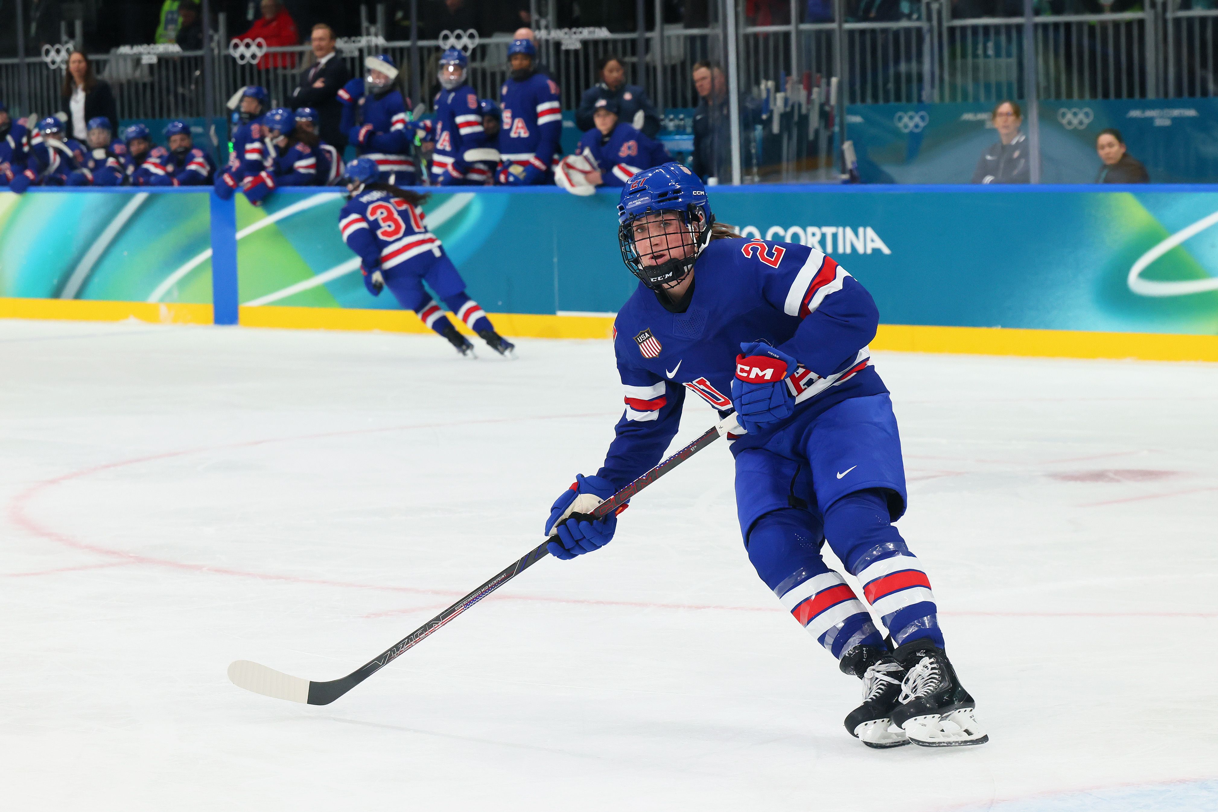 A USA women's ice hockey player in blue uniform with red and white stripes skates on ice holding a hockey stick during a game, with teammates and coaches watching from the bench.