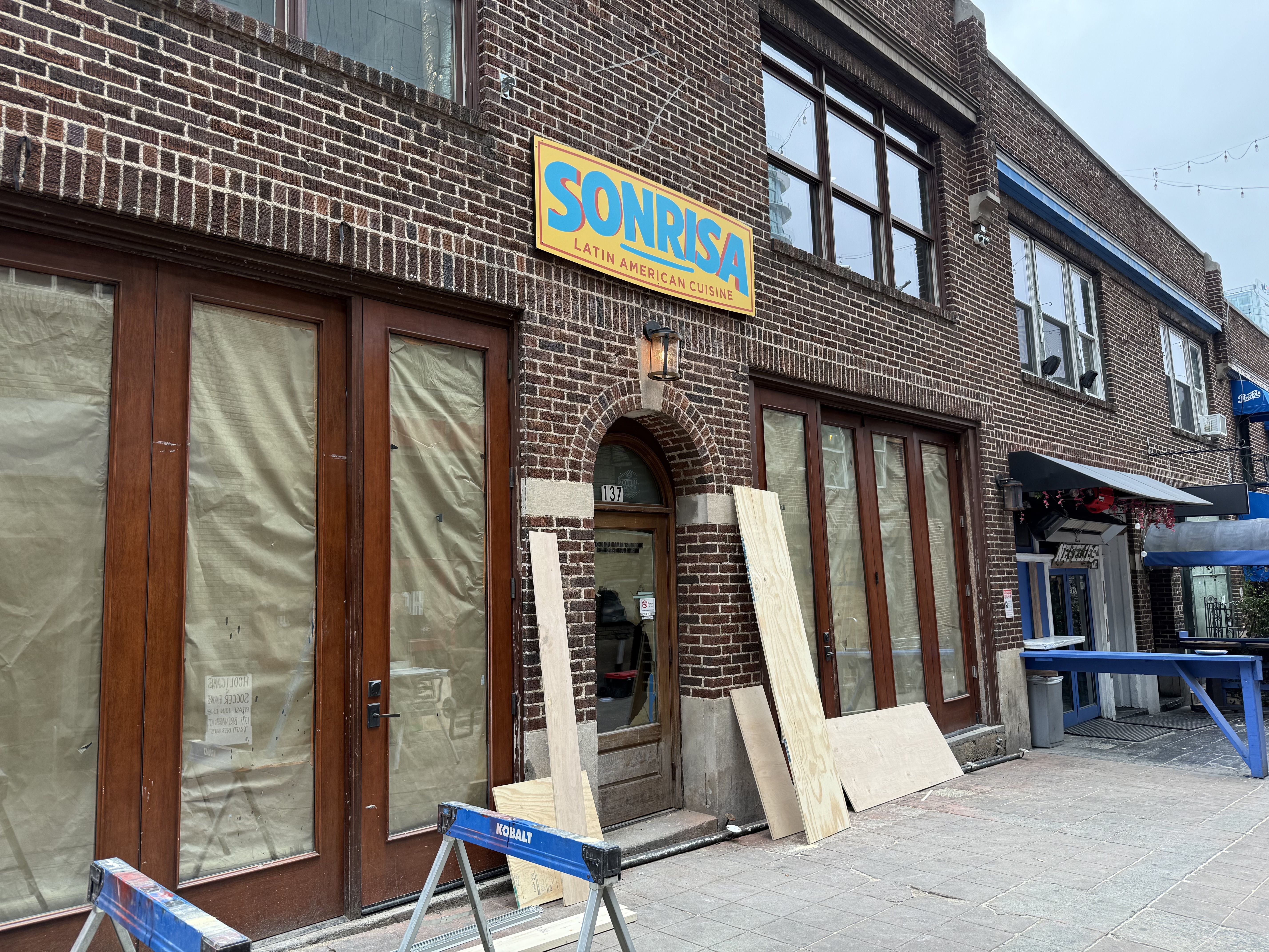 Exterior of a brick building with boarded-up windows and doors, featuring a colorful yellow and blue sign reading "SONRISA Latin American Cuisine" above the entrance, with construction materials nearby.