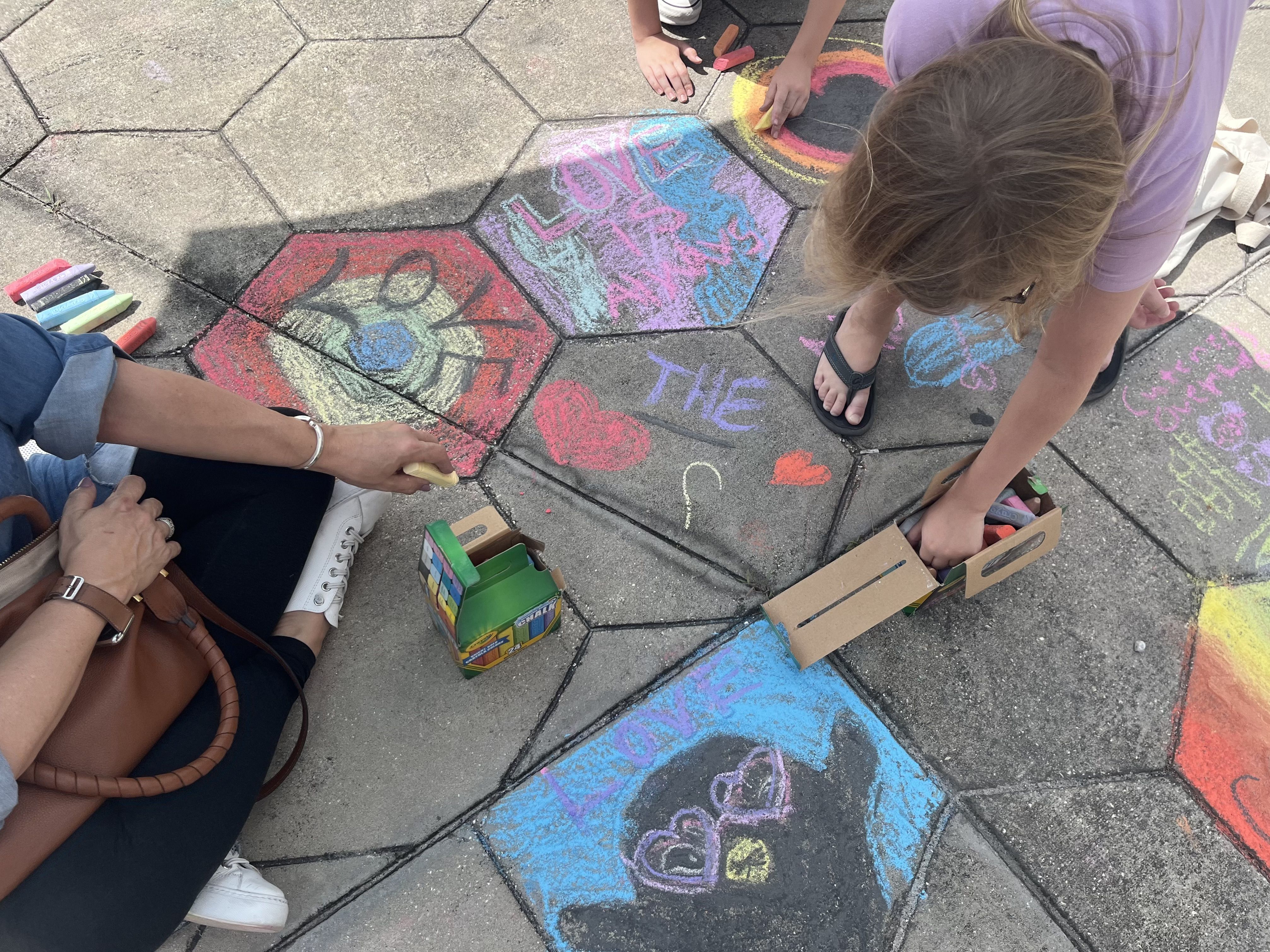A woman and a child draw rainbows and messages of love with sidewalk chalk. 