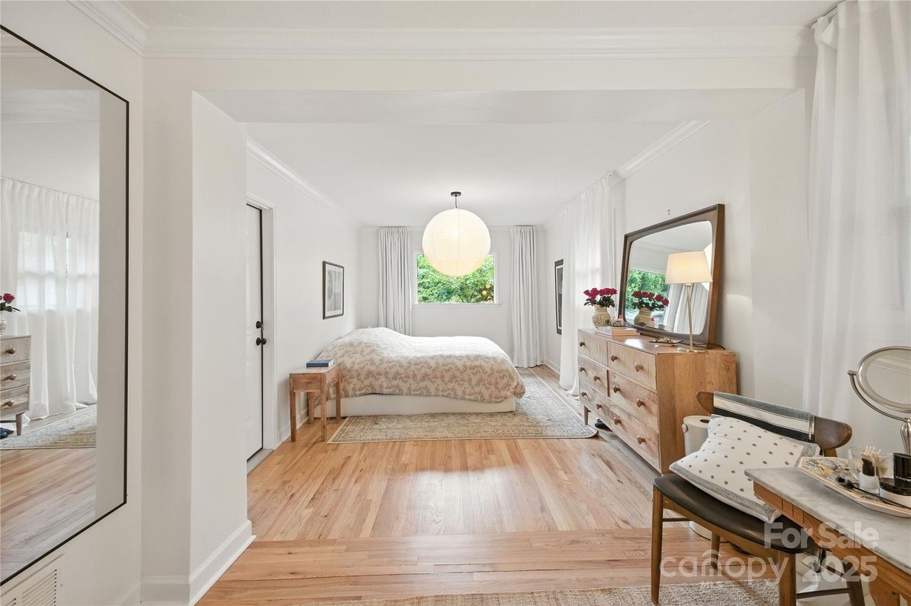 Bright bedroom with white walls, wooden floor, large paper lantern light, floral bedspread on bed, wooden dresser with mirror and red flowers, and a cozy chair with cushions.
