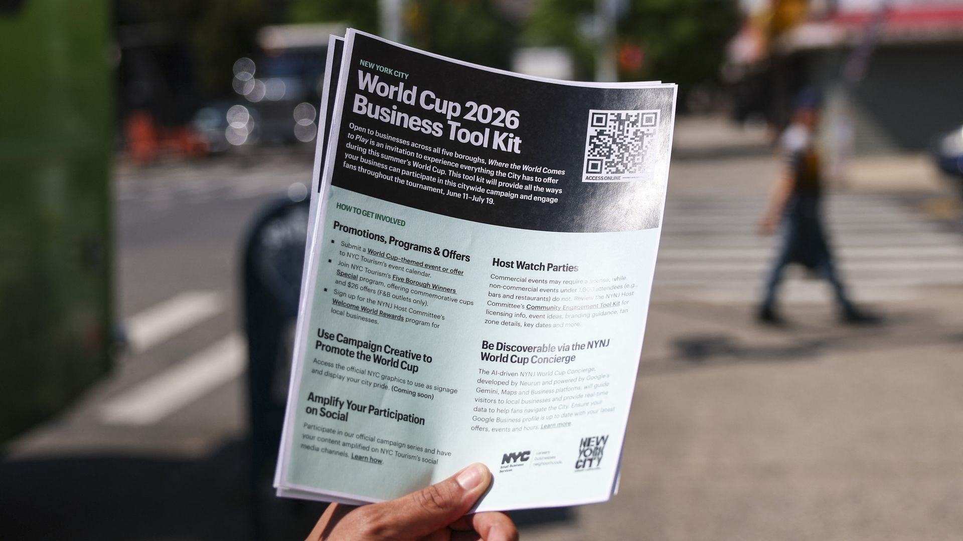 Hands hold a black-and-light-blue flyer titled "World Cup 2026 Business Tool Kit" with a QR code on the right, set against a blurred city street background.