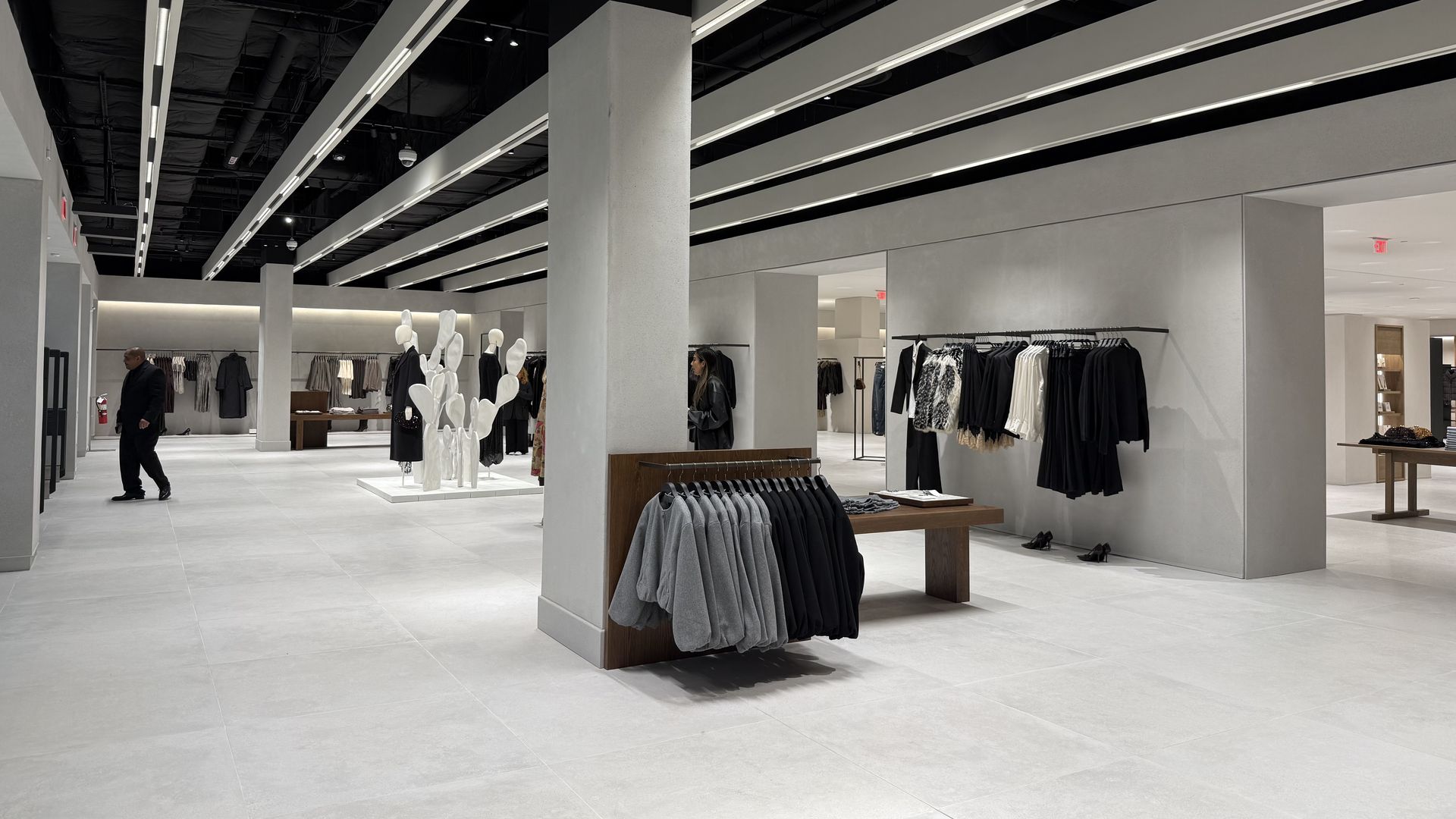Spacious modern clothing store with gray tiled floor, minimalist design, black and gray clothing racks, mannequins, and ceiling lights with exposed black ductwork.