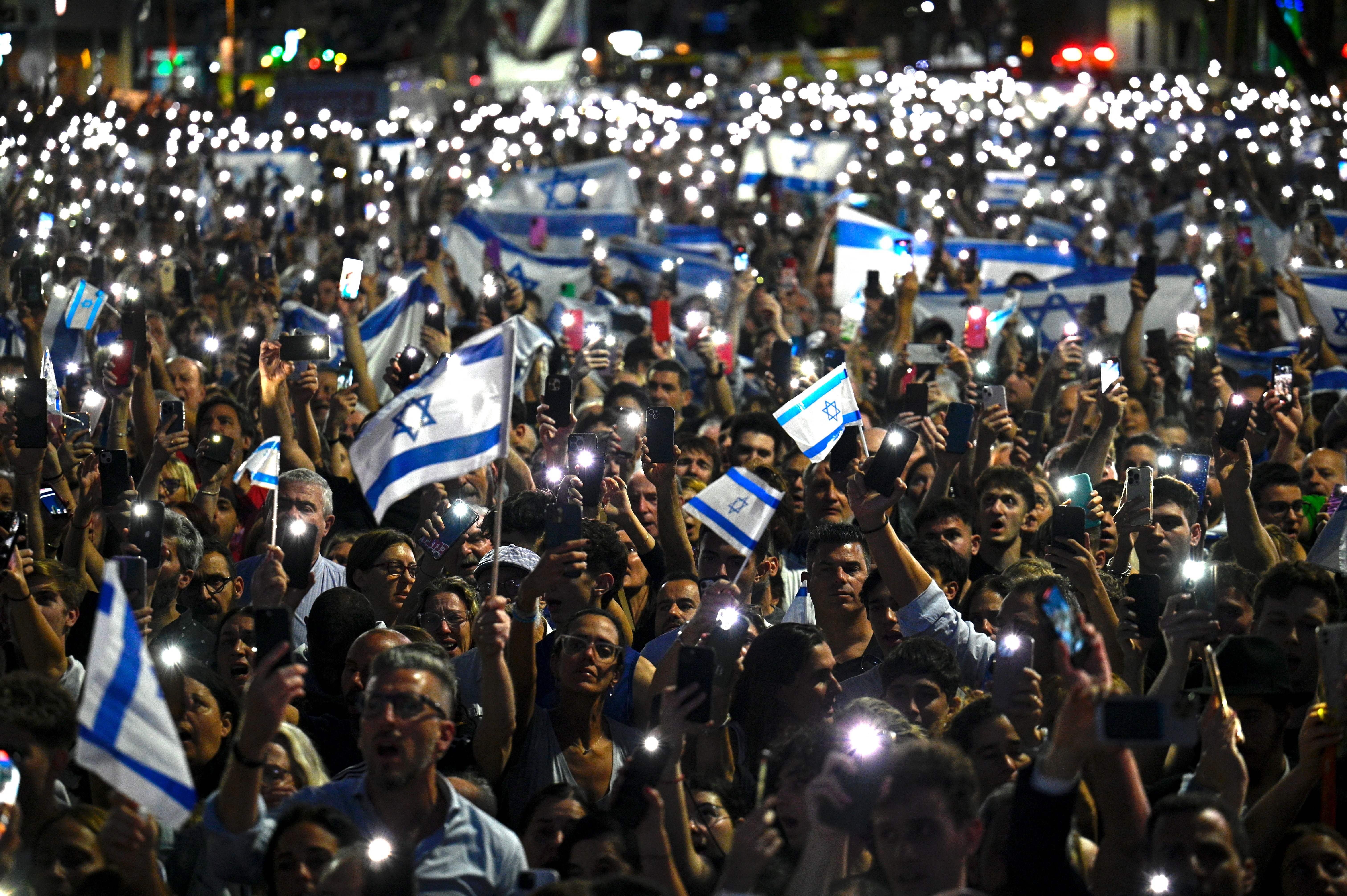 A wide shot of a crowd of protestors holding up lit mobile phones and Israeli flags. 