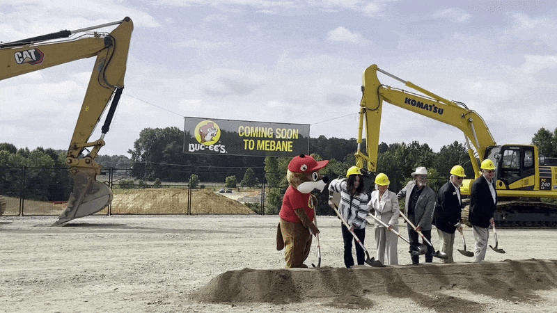 Gif of people in hard hats and one wearing beaver costume shoveling dirt at a ceremonial groundbreaking. Behind them are excavators and a sign that says "Buc-ee's COMING SOON TO MEBANE."