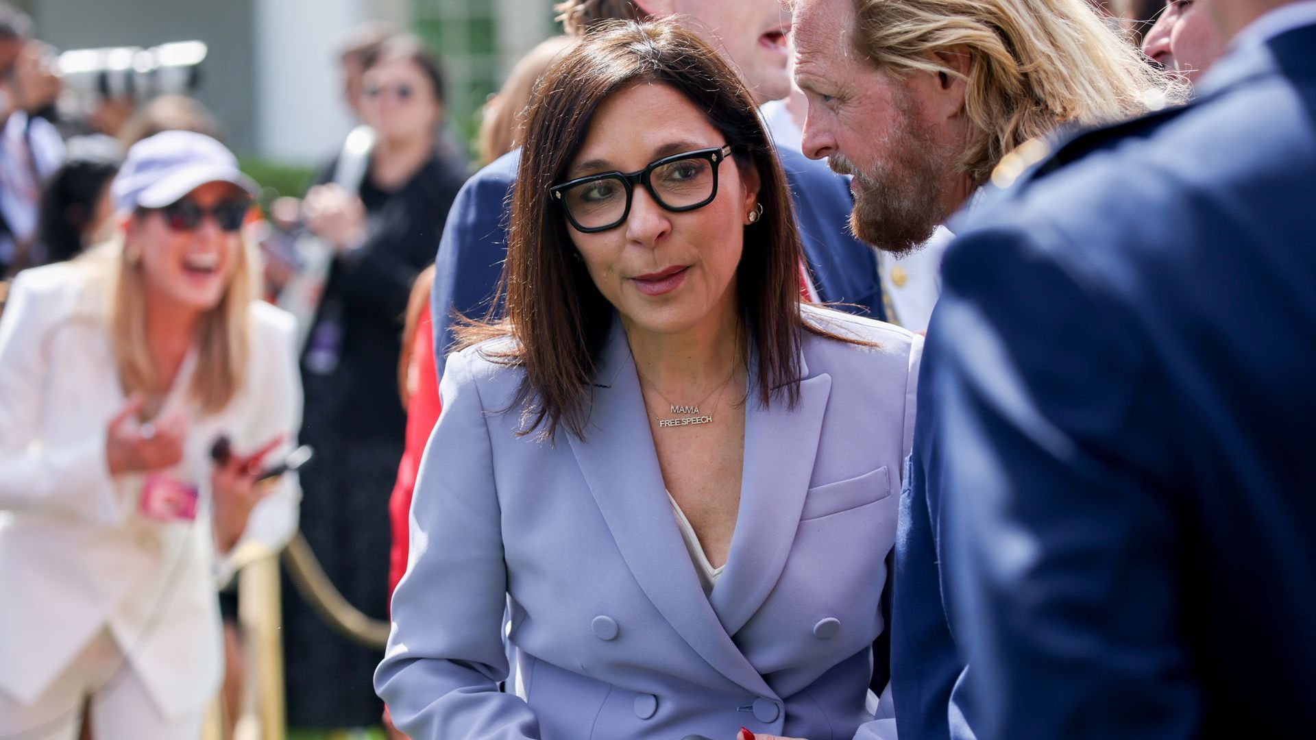 Woman in light purple blazer and glasses talking to a man with long blond hair and beard. Background shows people dressed up outdoors, one woman laughing in white outfit and cap.