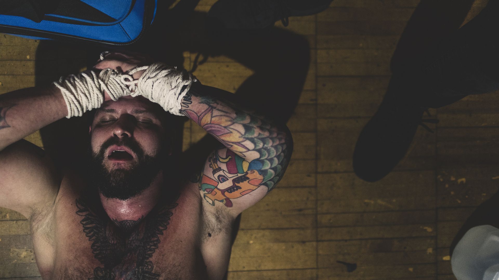 Top-down view of a shirtless, bearded man with colorful tattoos; wrists bound in white rope above his head on a wooden floor, with a blue bag nearby and dark shadows around.