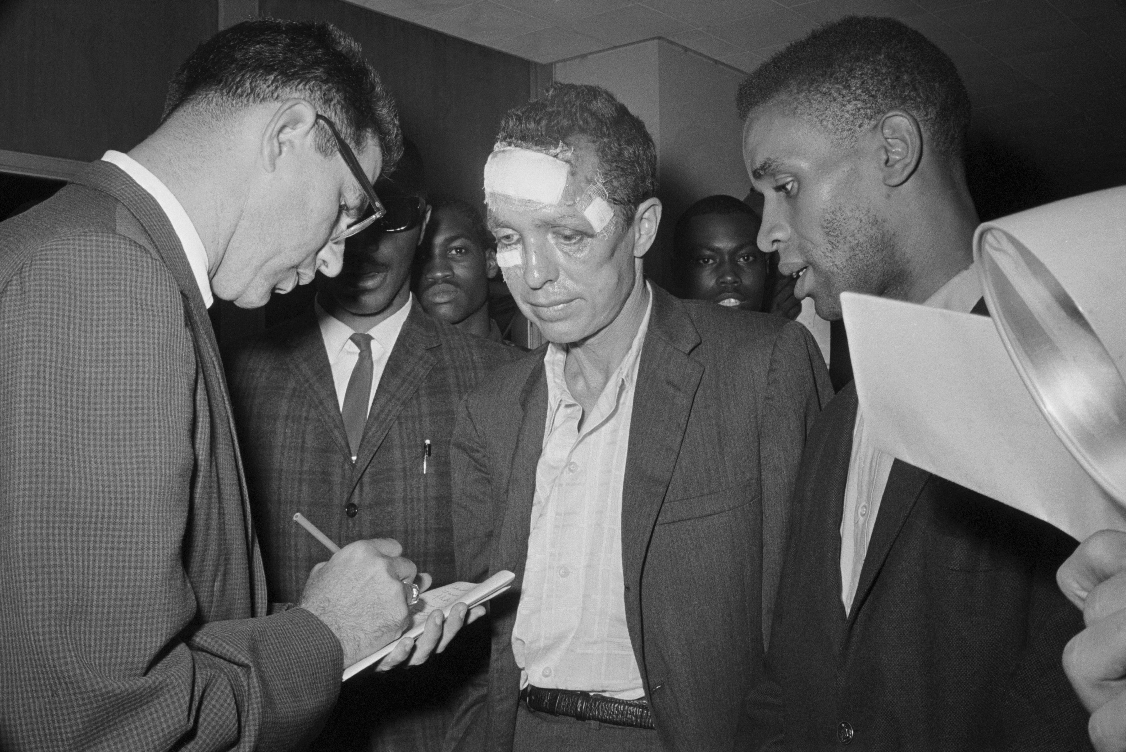 A black and white photo of a man interviewing two other people. One of the interviewees has bandages on his face.