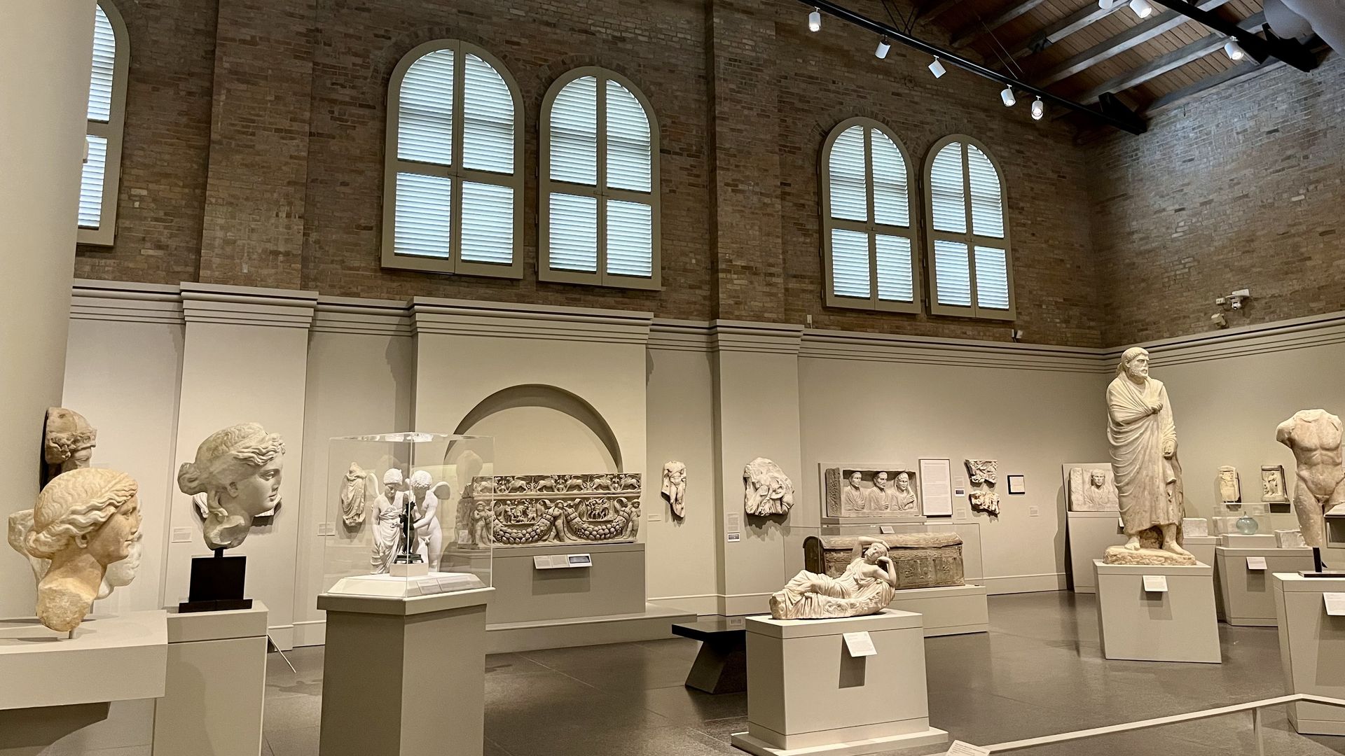 A museum gallery with high brick walls and arched windows, displaying ancient Greco-Roman sculptures and marble reliefs under soft lighting. Statues and busts are arranged on pedestals throughout the spacious, modern room.