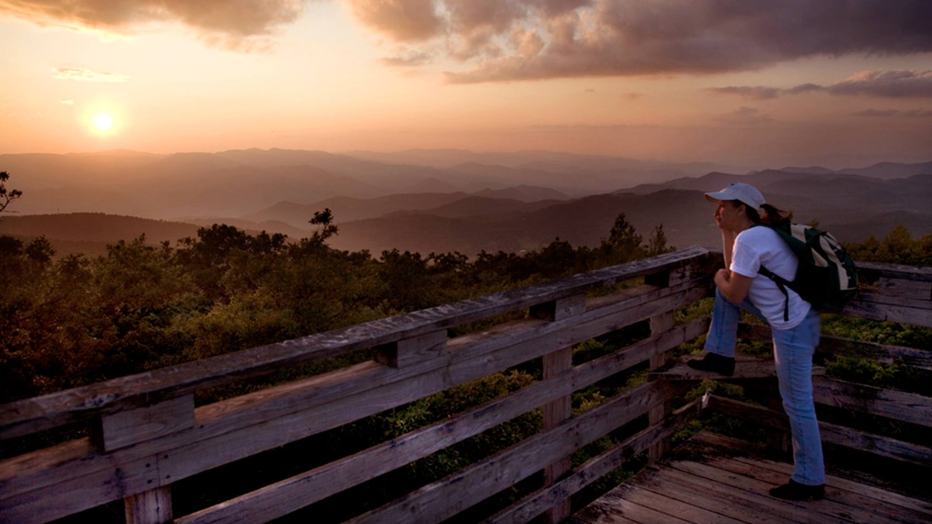An individual wearing a backpack rests their foot against a wooden railing and gazes at a vast rolling mountainscape 