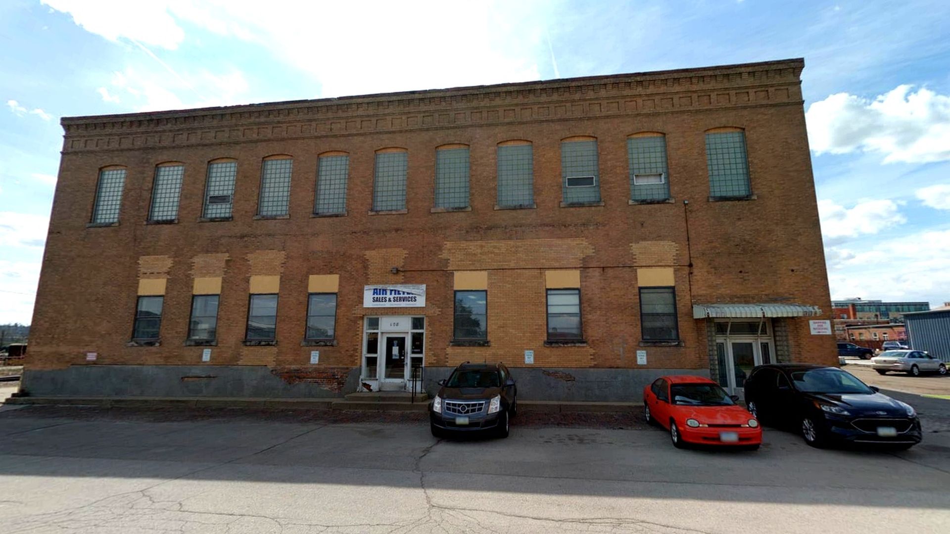 Two-story brick building with tall grid windows, a central door under a 'Sales & Services' sign, and three cars (gray SUV, red convertible, black sedan) parked in front under a blue sky.