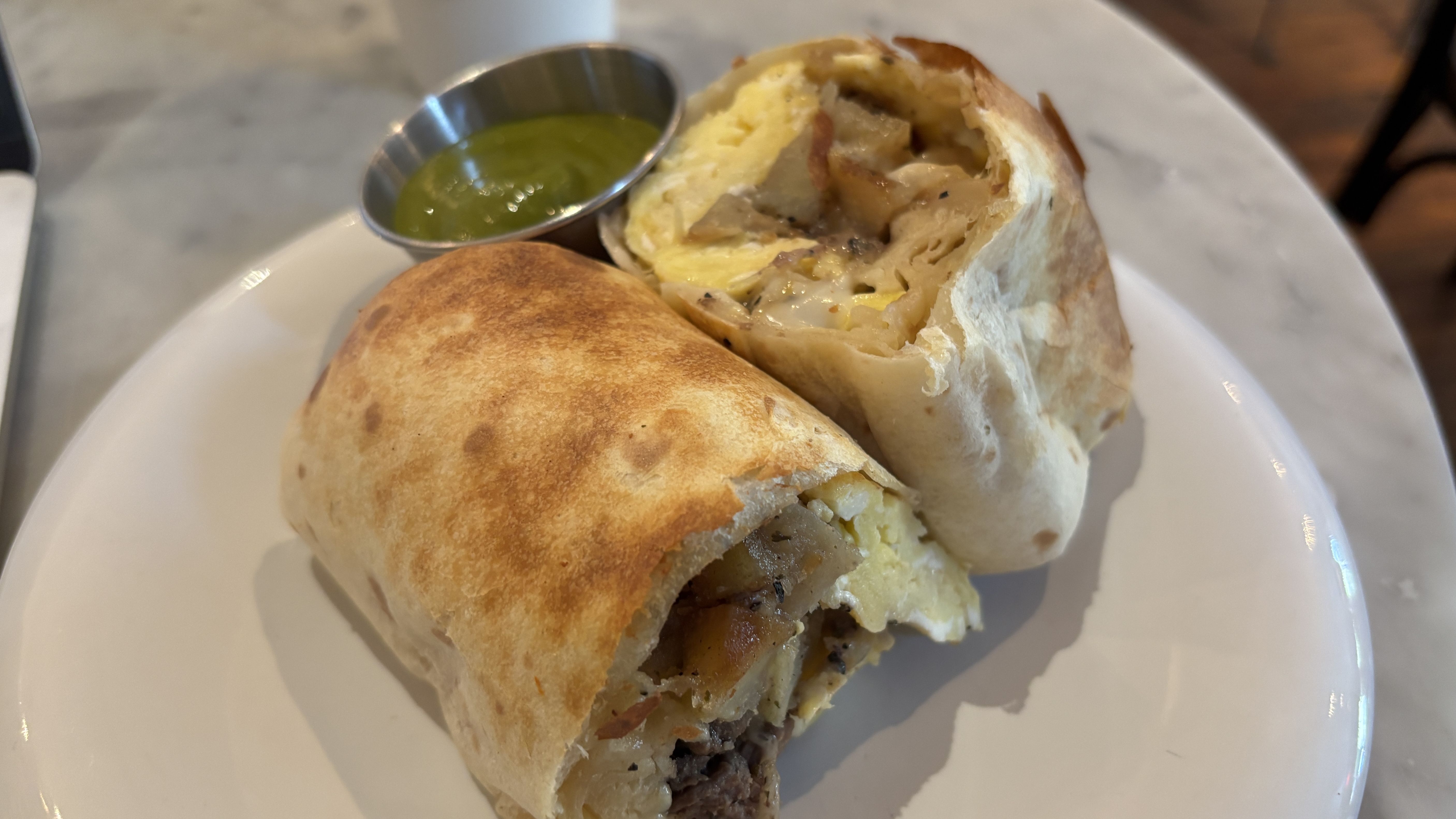 Toasted burrito cut open on a white plate, revealing scrambled eggs, potatoes, melted cheese and beef inside; a small metal cup of green salsa sits beside it.