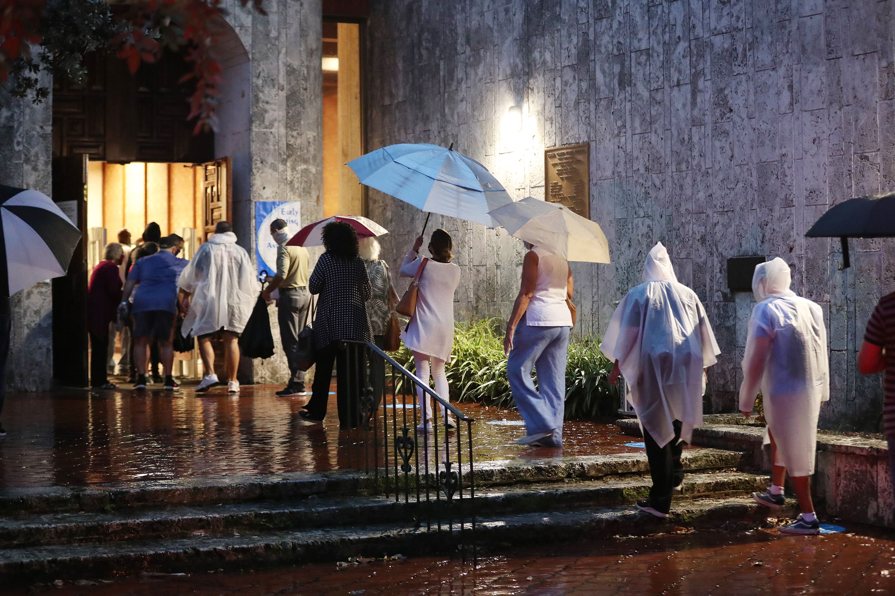 A line of people stand in the rain, wearing ponchos and holding umbrellas, to vote at night