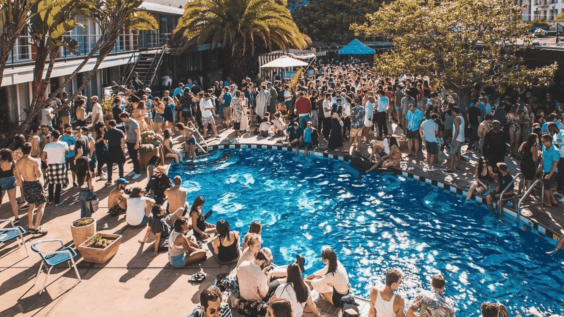 Crowded outdoor pool party with many people sitting by and standing around a bright blue pool, surrounded by palm trees, buildings, and clear sky.