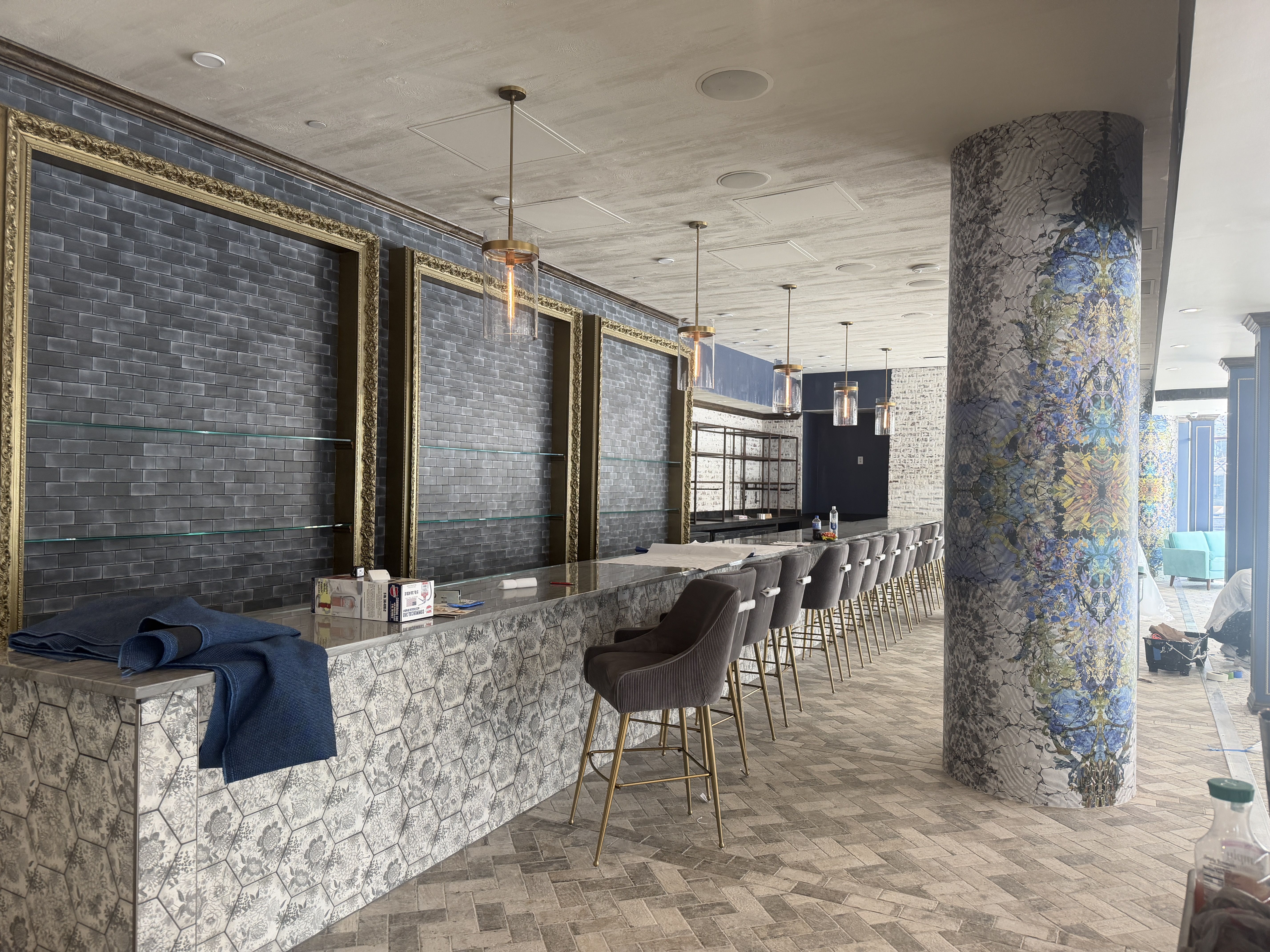 Long bar with hexagonal patterned tiles, dark gray cushioned high chairs with gold legs, hanging pendant lights, dark blue brick wall with gold frames, and decorative pillar with blue and yellow pattern.
