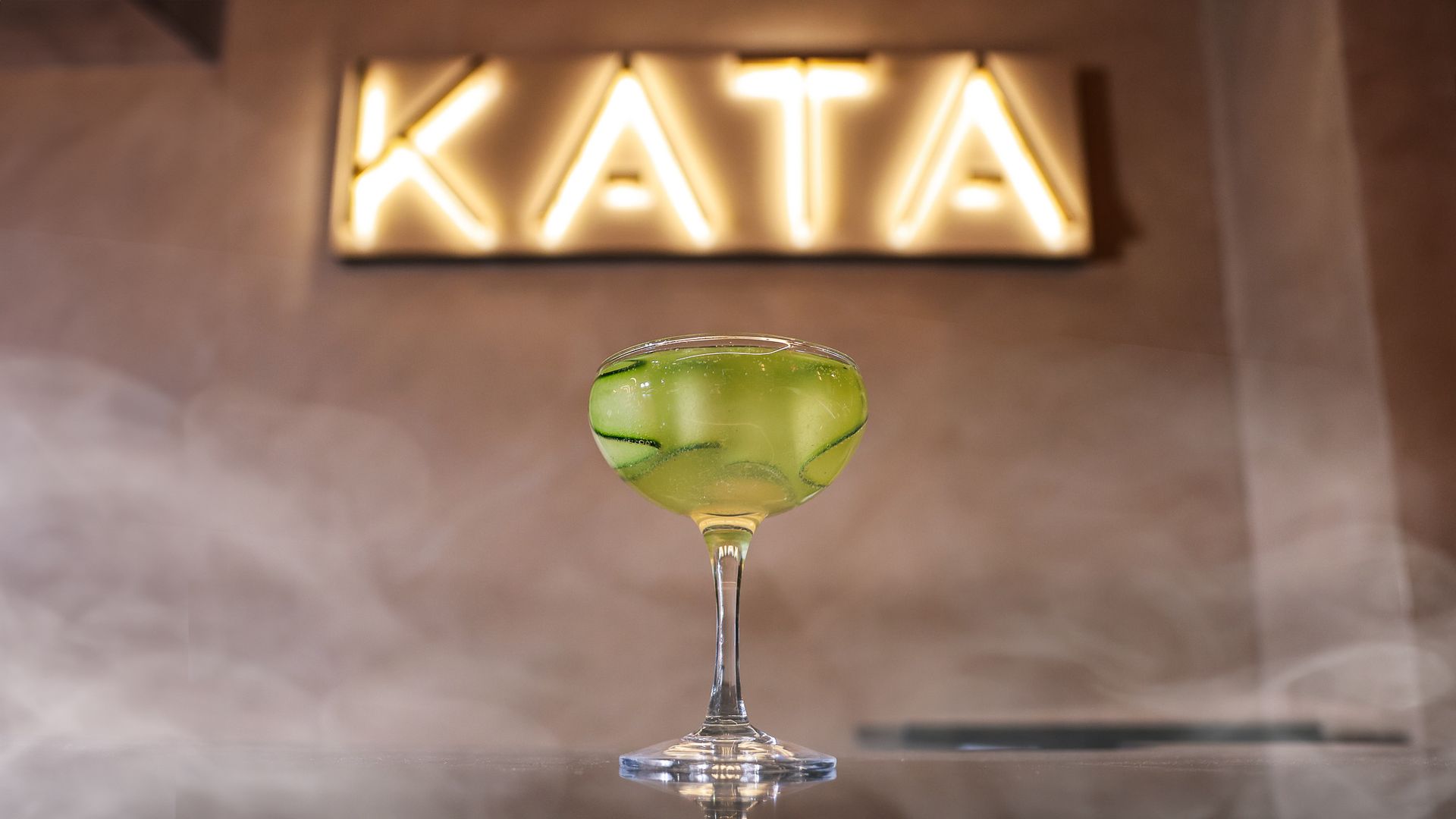 A cucumber cocktail on a bar with a glowing "KATA" sign behind