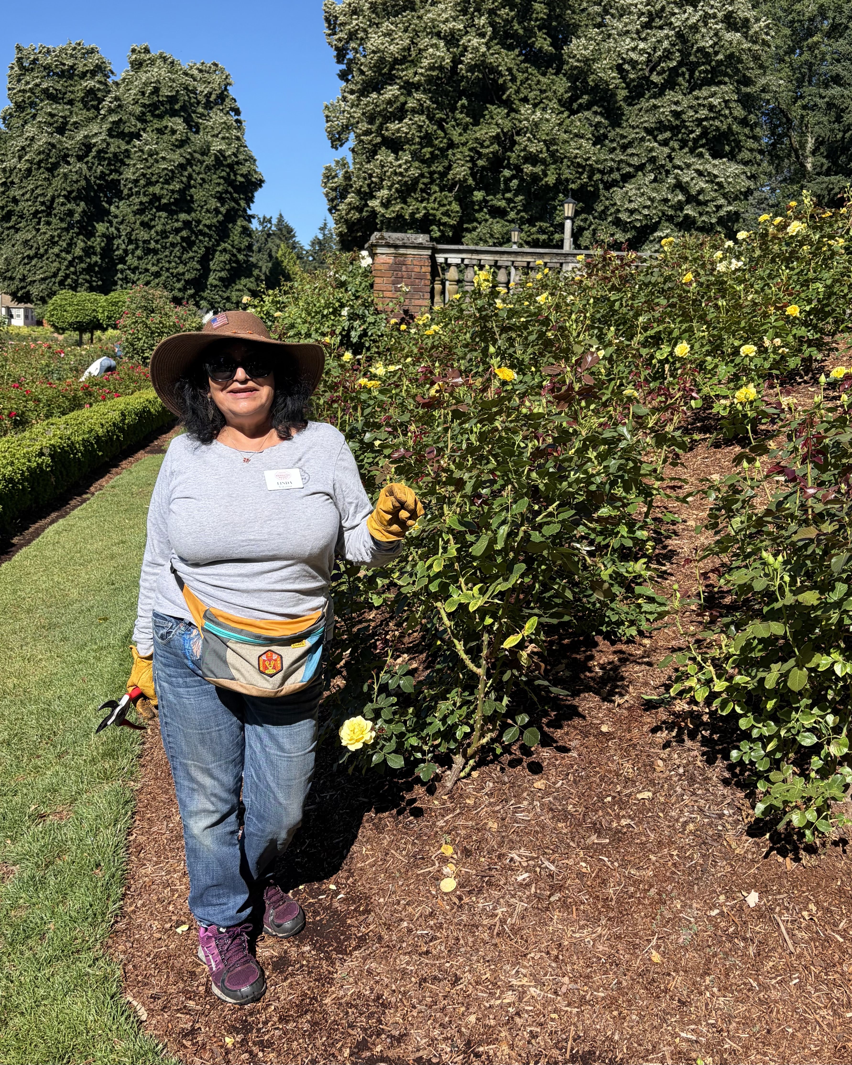 Smiling woman in a wide-brim hat and sunglasses stands in a rose garden with yellow blooms, wearing a gray shirt, jeans, and a beige waist bag; a brick balustrade and tall trees rise behind her.