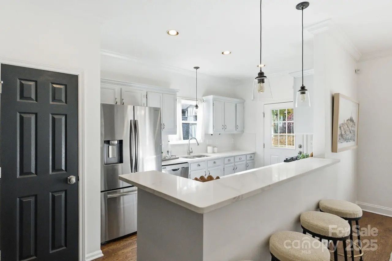 Bright white kitchen with white cabinetry, stainless steel refrigerator, black door, three hanging pendant lights, white countertop bar, and three round beige stools.