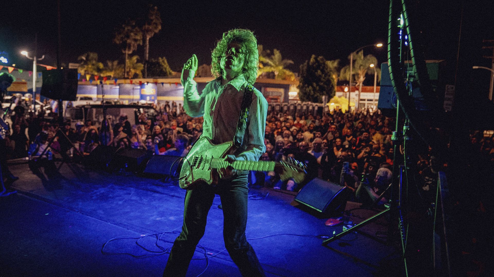 Nighttime concert: a guitarist with long hair and a white electric guitar, bathed in green stage light, performing on a blue stage before a large crowd at an outdoor festival with pennant banners.